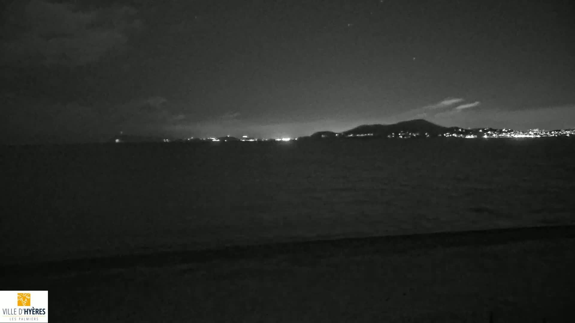 A nighttime view of a dark body of water with a distant, faintly lit cityscape under a mostly cloudy sky.