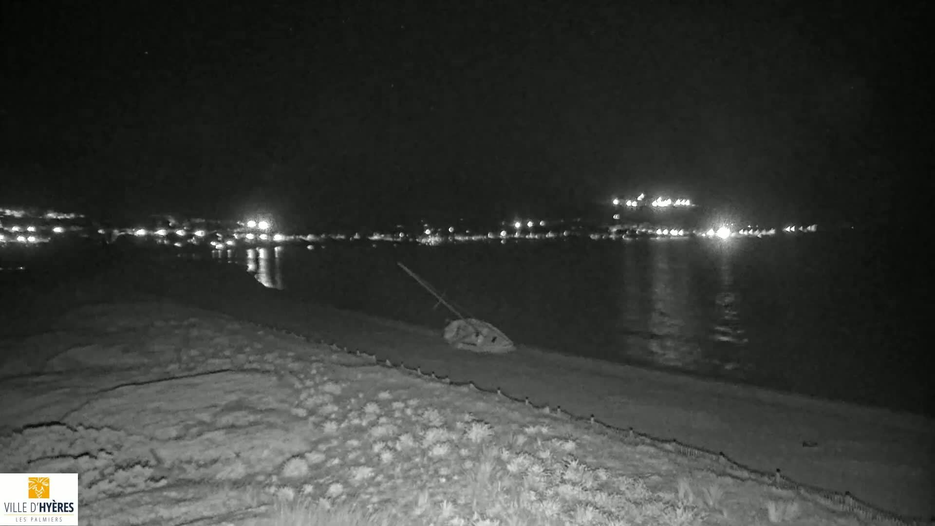 A capsized sailboat lies on a dark beach at night, with lights from a town visible across the water.