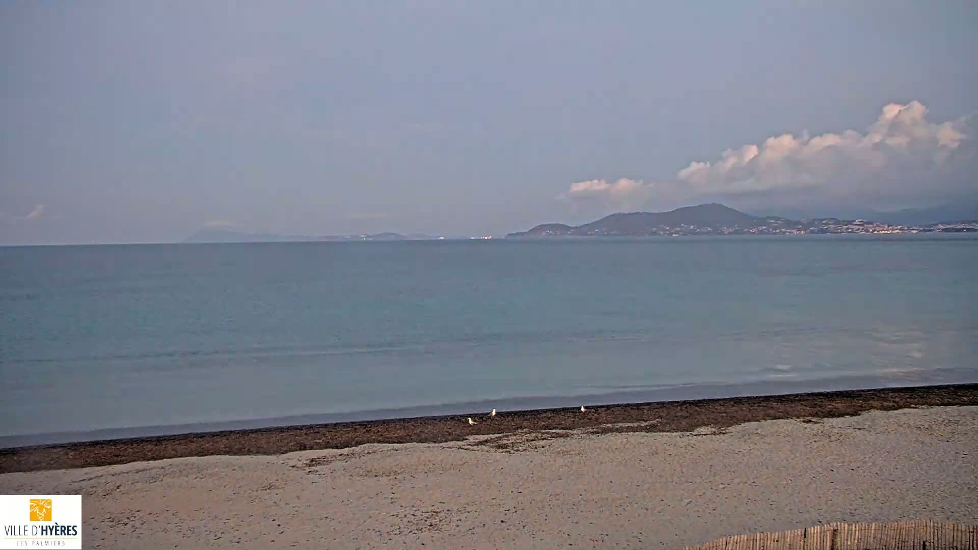 A sandy beach with a few birds on the shore overlooks a calm sea and distant mountains under a partly cloudy sky.