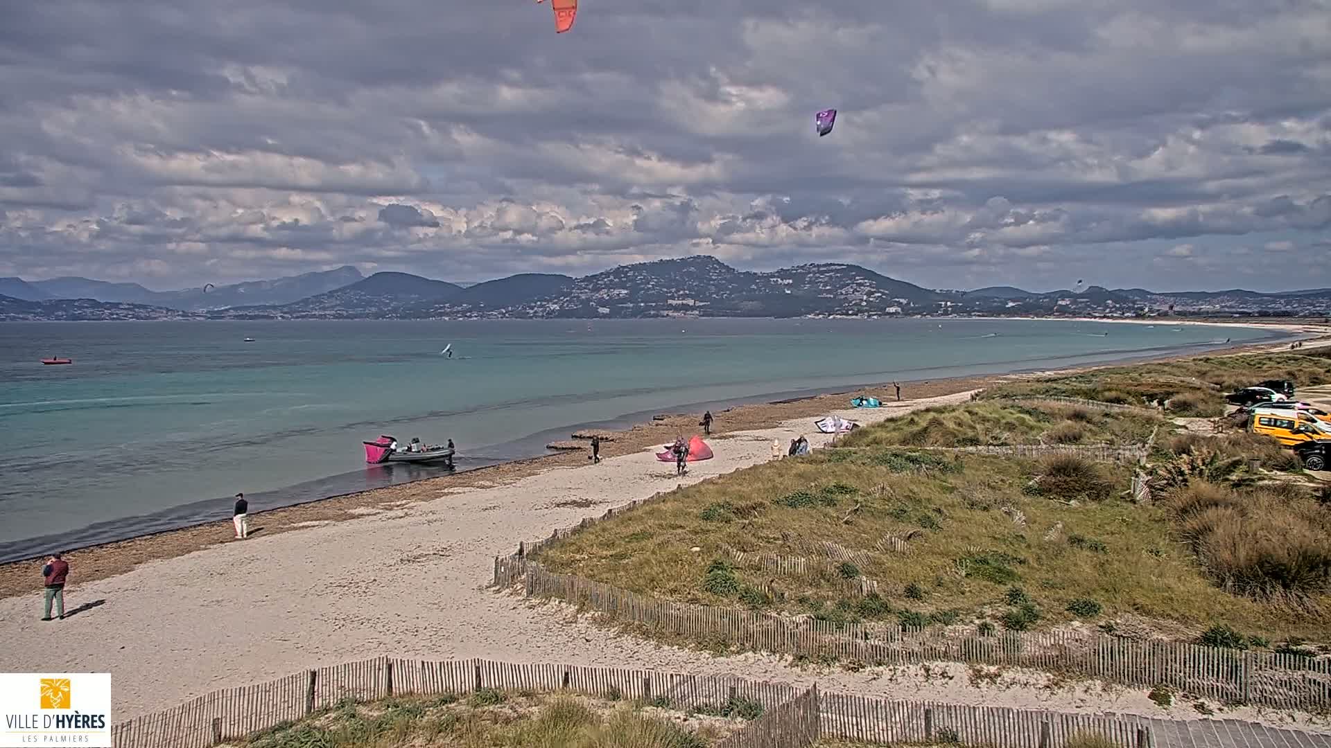 A sandy beach with several people and kite surfers, a few small boats, and a line of parked vehicles is visible under a partly cloudy sky, with mountains in the distance.
