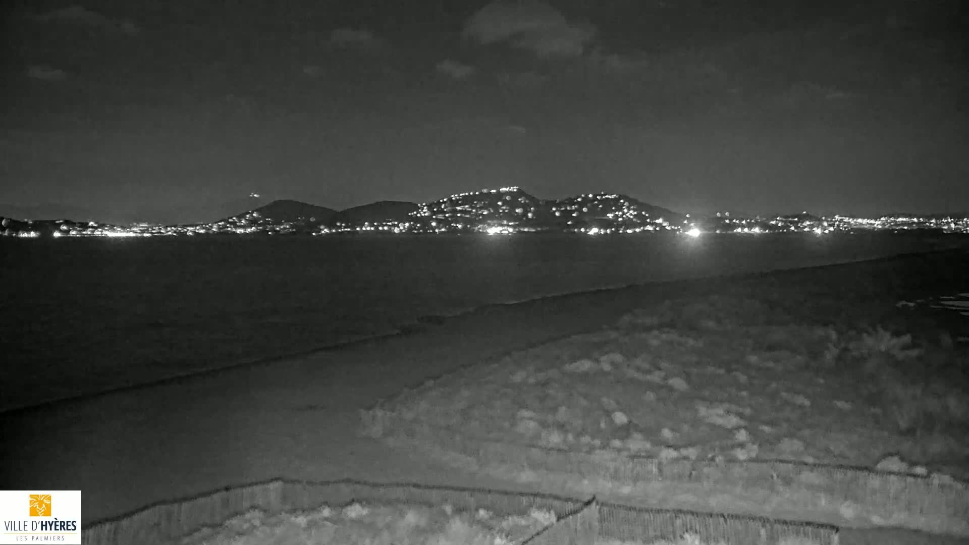 A nighttime view of a dark, calm body of water with a hill dotted with lights in the distance under a cloudy sky.