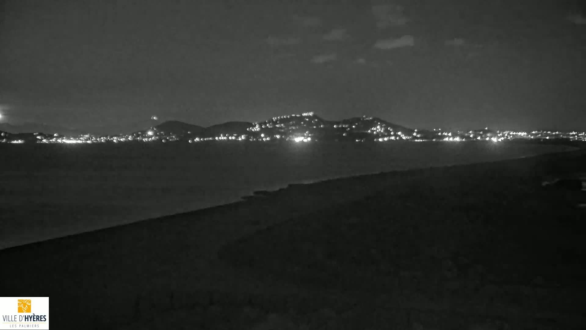 A nighttime coastal scene shows a dark shoreline in the foreground with a distant town illuminated by lights across a dark body of water under a mostly clear sky.