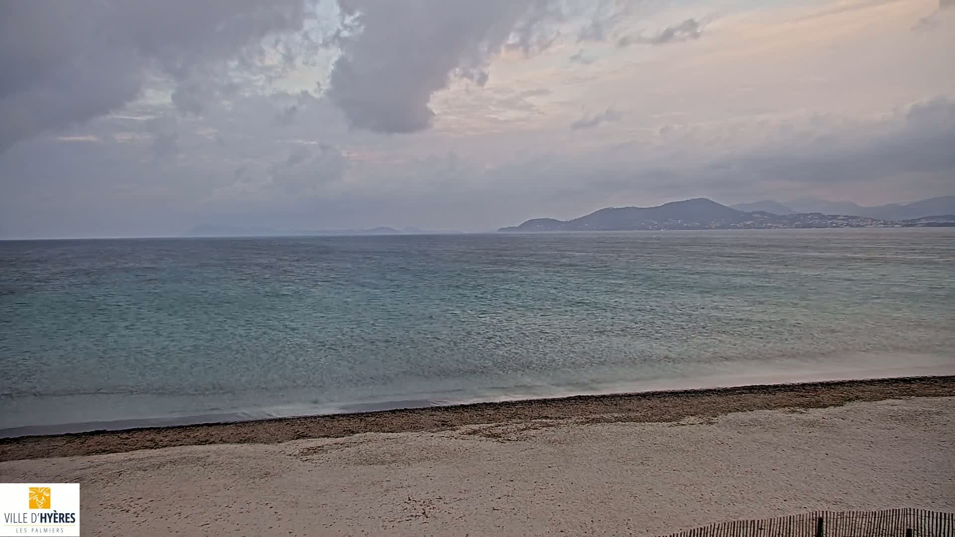 A sandy beach meets a calm, teal sea under a cloudy sky, with distant mountains visible on the horizon.