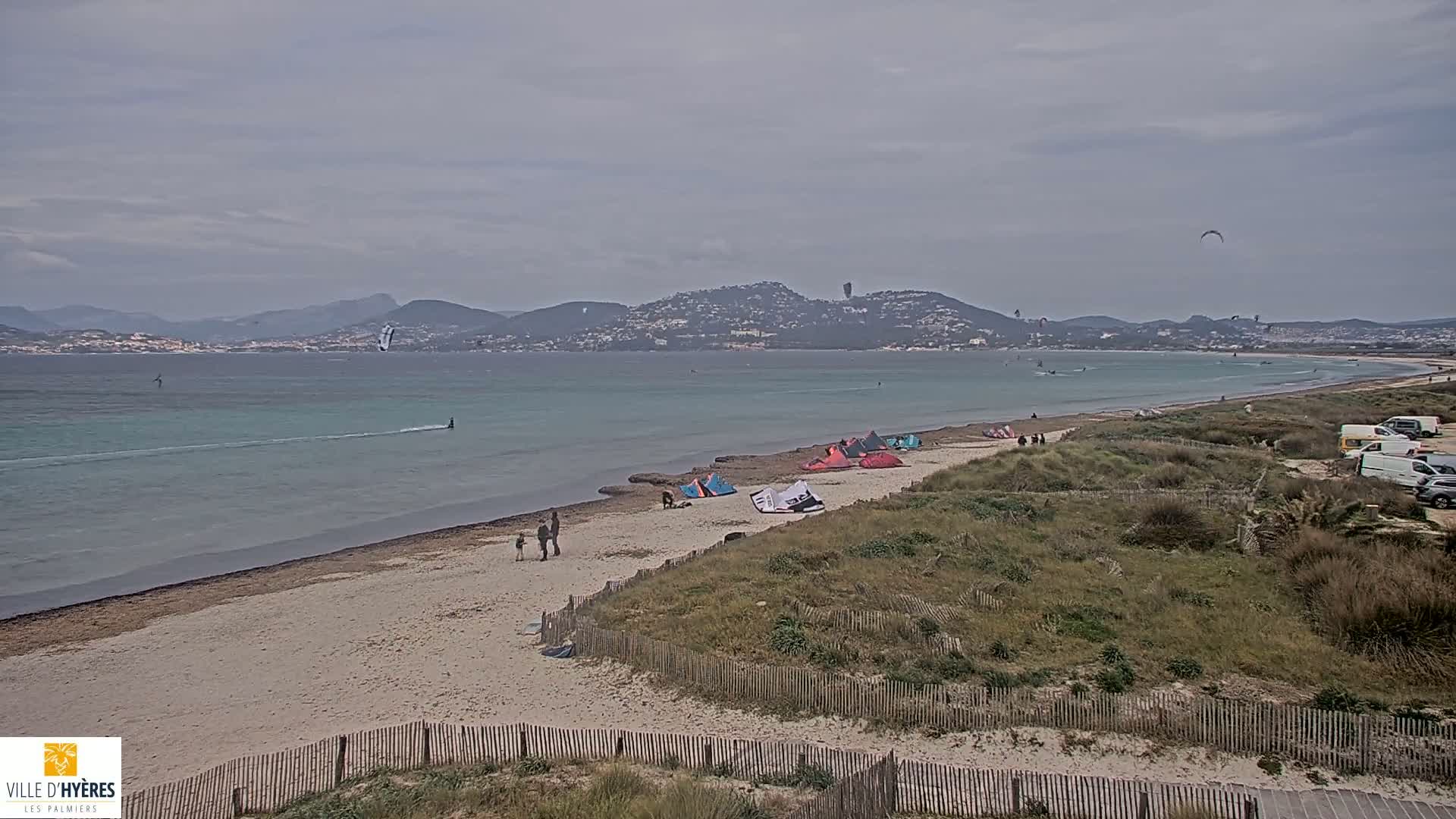 A sandy beach with several kitesurfers in the water and on the shore, under an overcast sky, and with mountains in the distance.