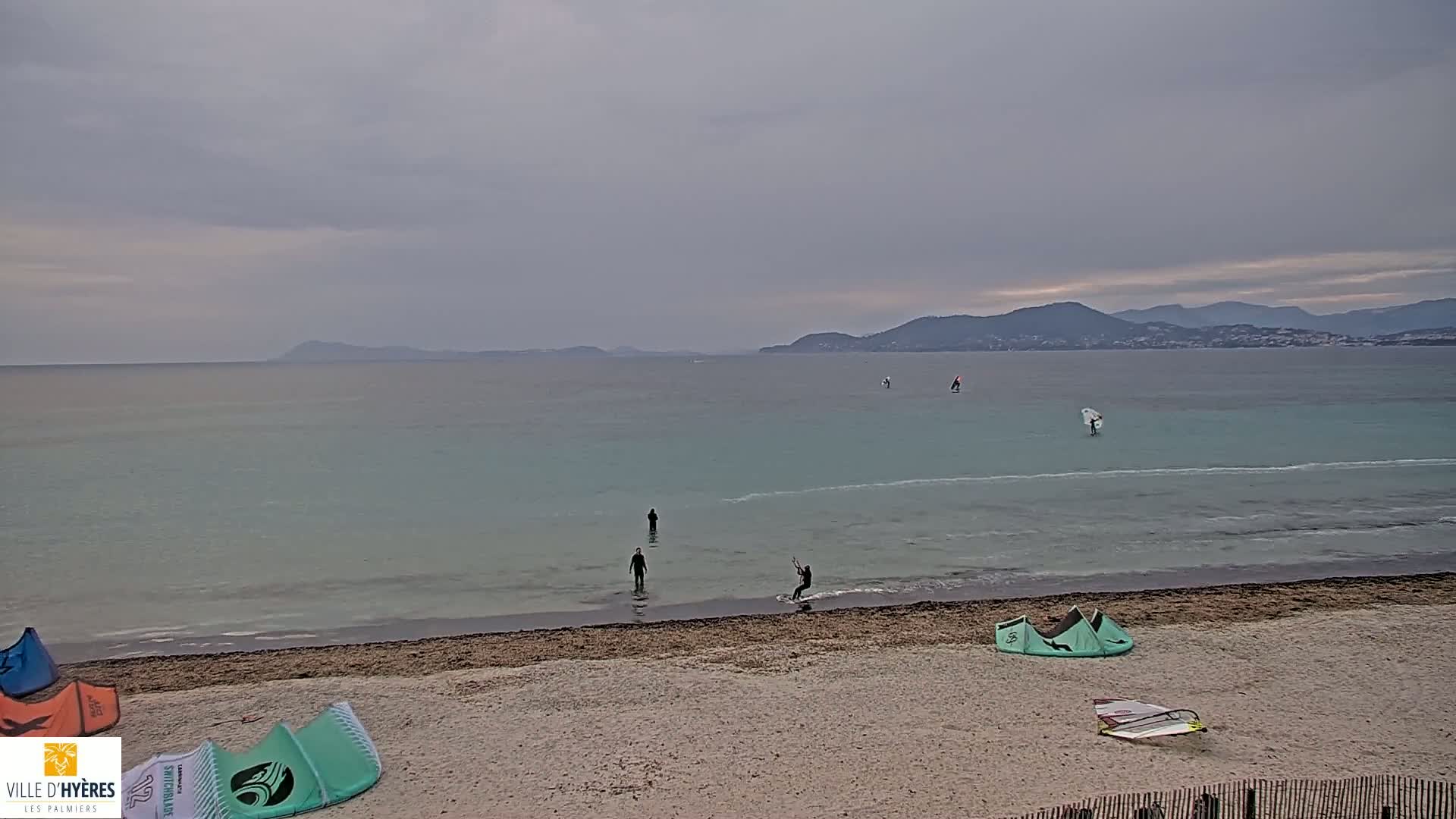 A sandy beach with several people windsurfing and kitesurfing in calm, pale turquoise water under an overcast sky, with distant mountains visible on the horizon.