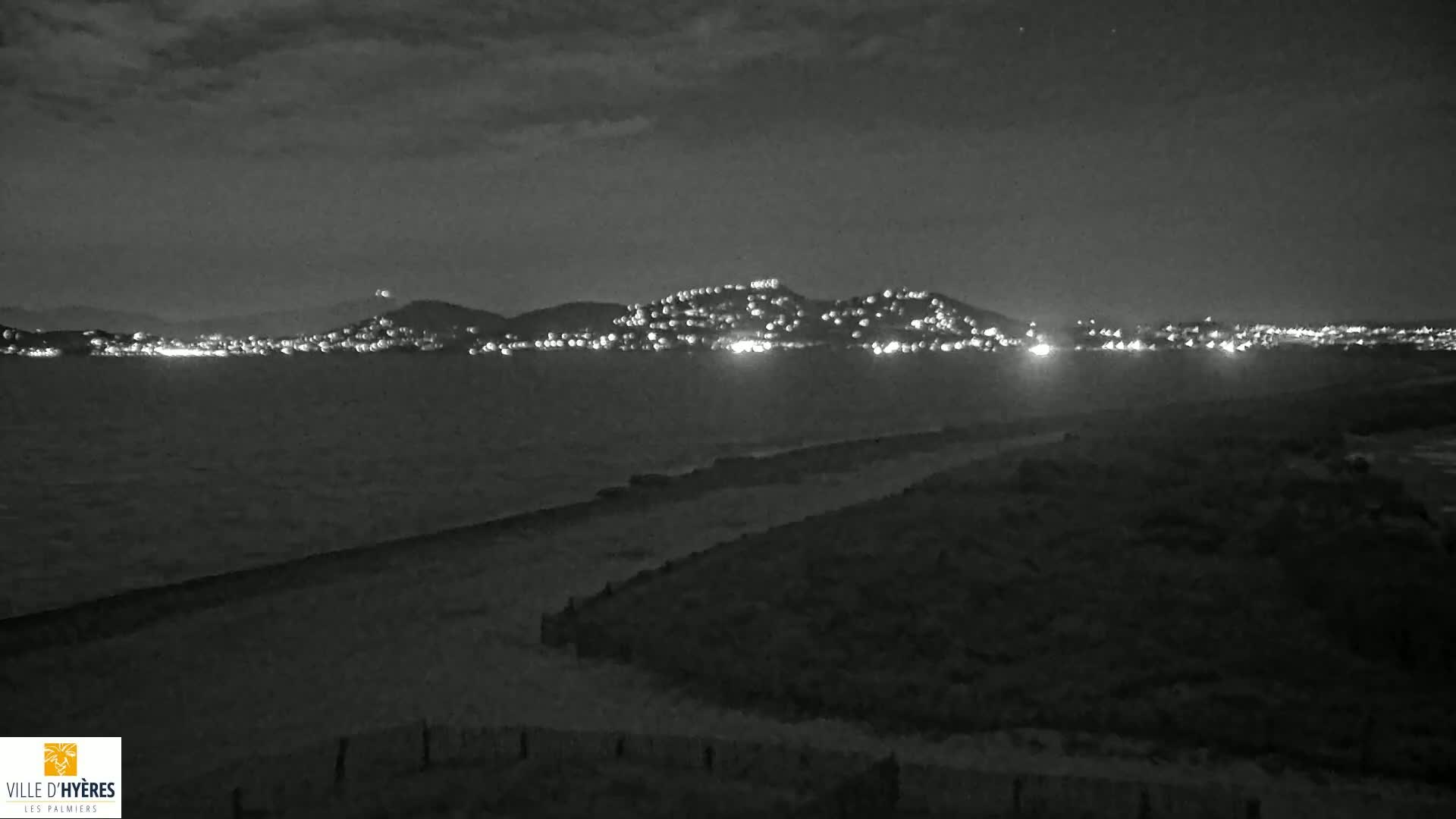 A nighttime, grayscale view shows a dark beach and a body of water with a distant city illuminated on a hill under a cloudy sky.