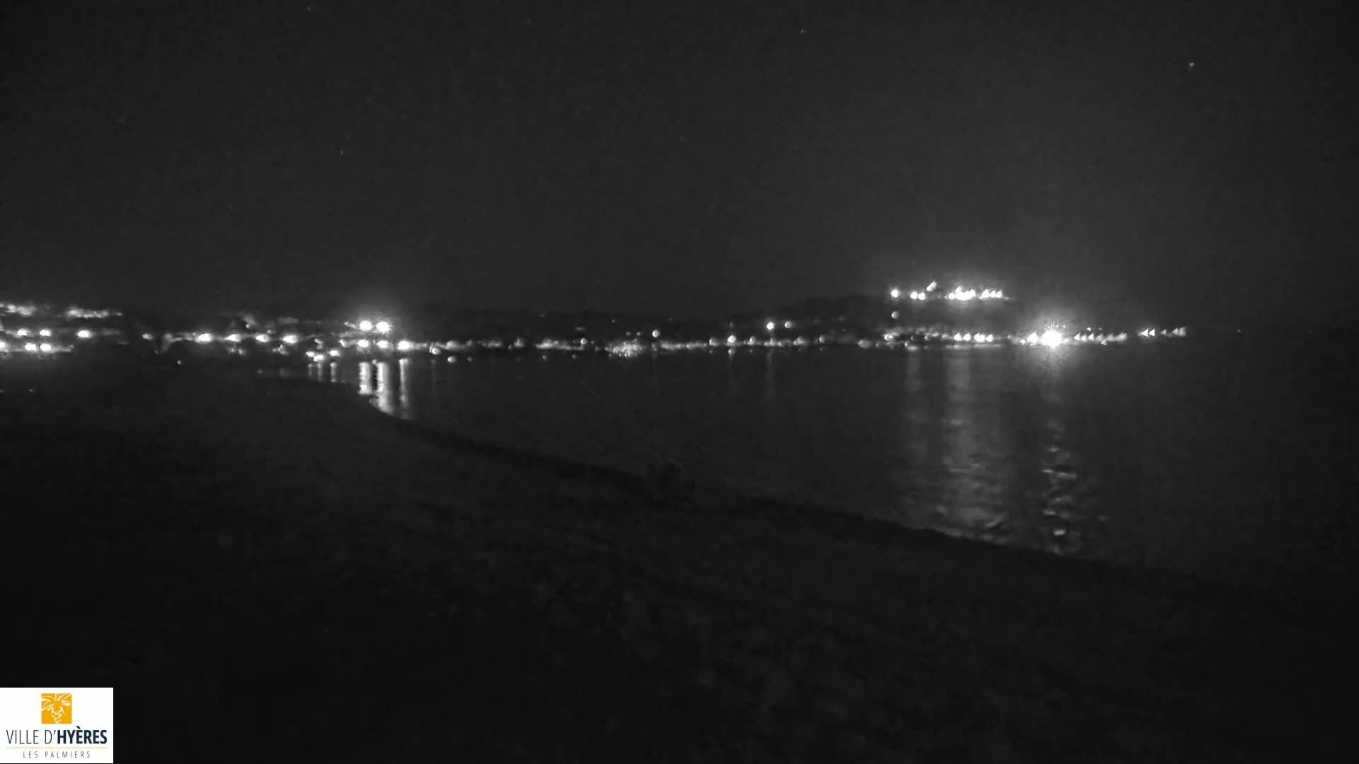 A dark, nighttime beach scene shows a calm body of water reflecting the lights of a town across the bay under a clear sky.
