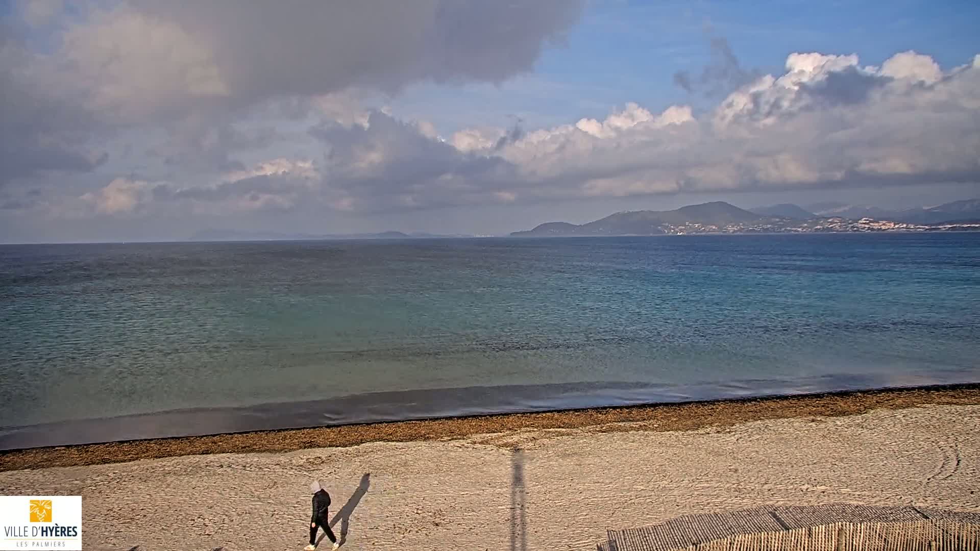 A person walks on a sandy beach next to a calm, blue-green sea under a partly cloudy sky with distant mountains visible on the horizon.