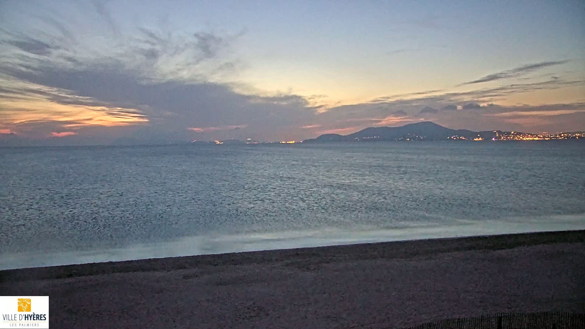 A dark beach meets a calm sea under a twilight sky, with distant lights illuminating a mountain range across the water.