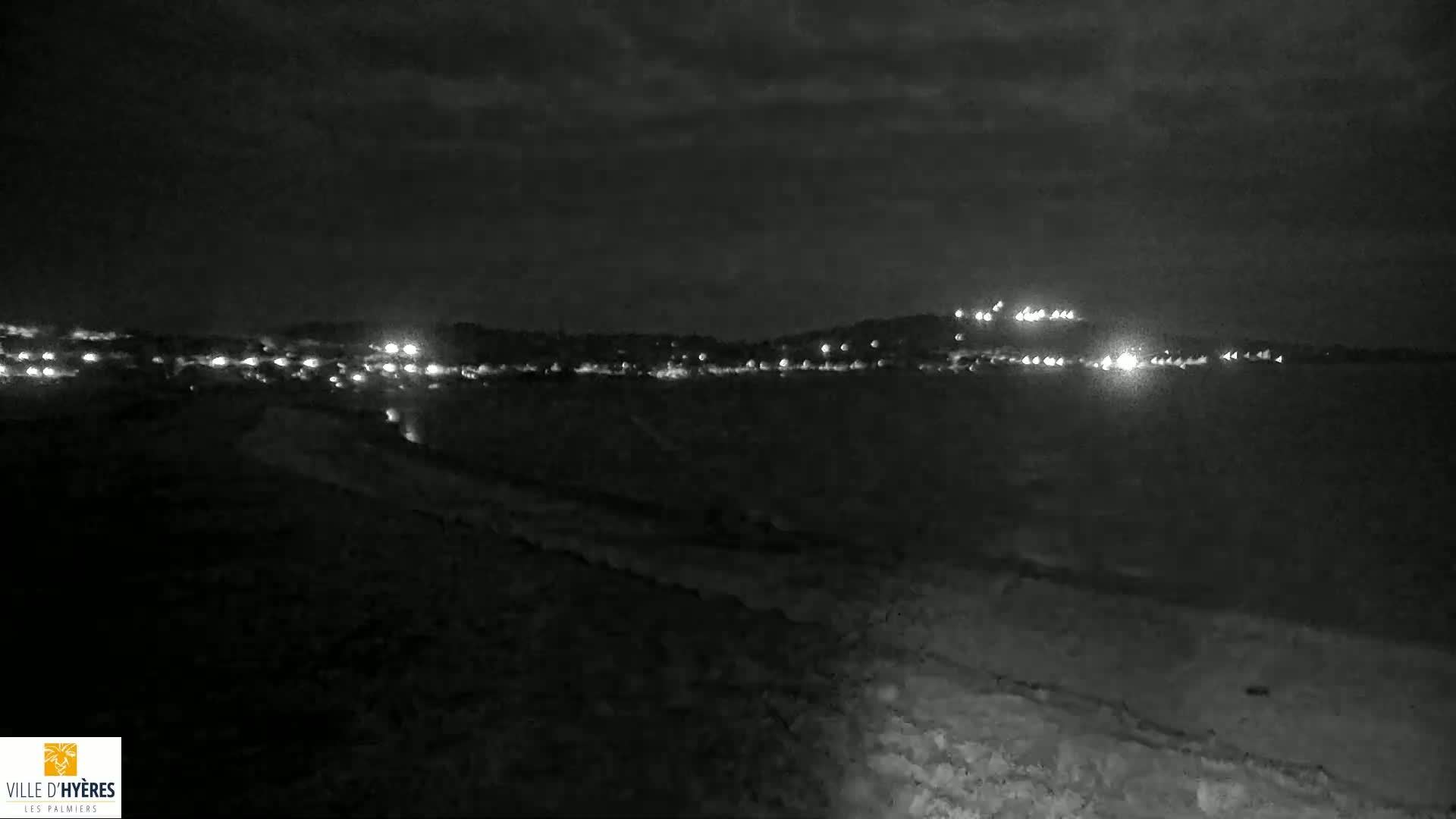 A nighttime, dark-grey-toned view of a distant town's lights across a body of water, under a cloudy sky.