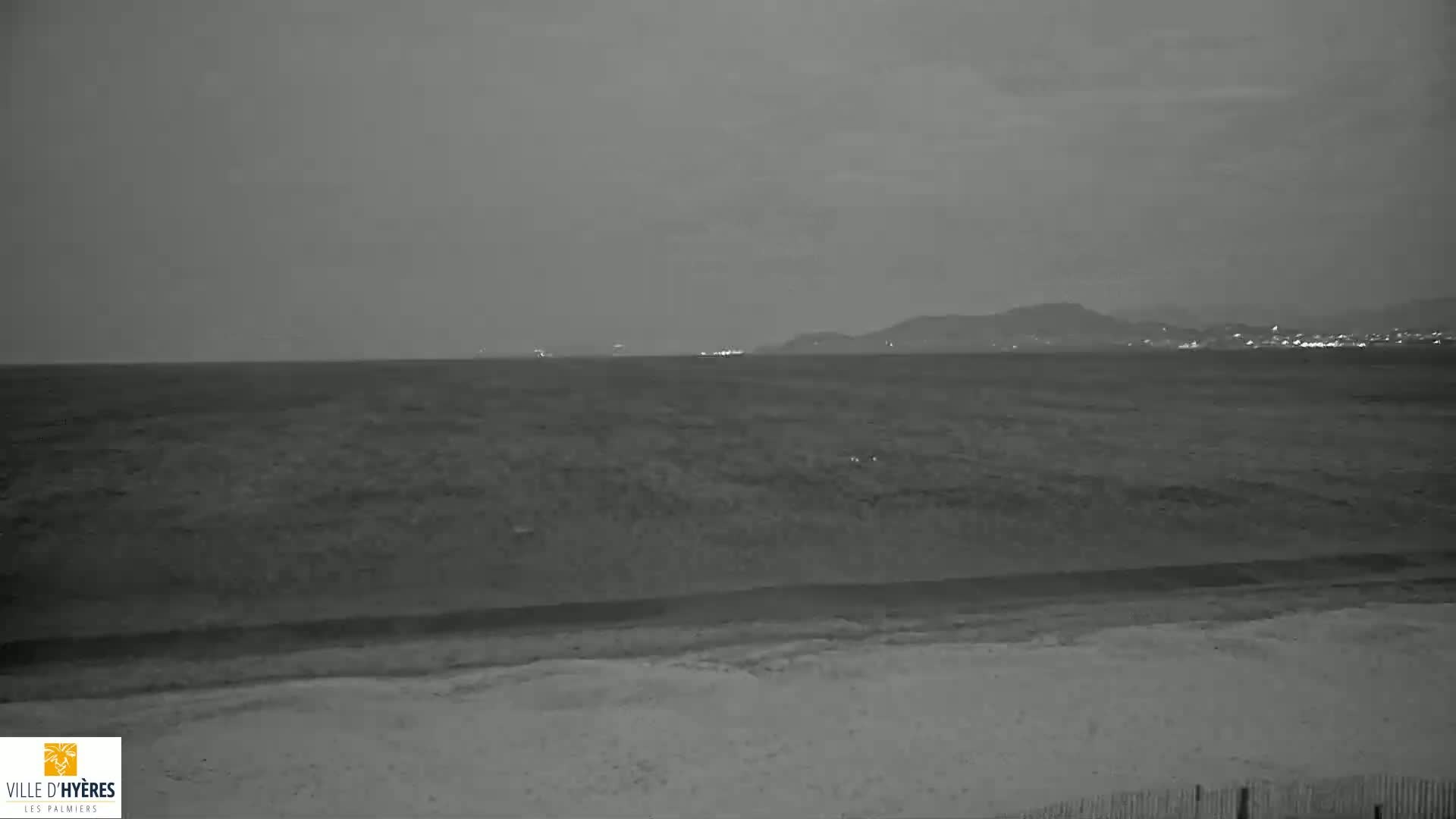 A dark, nighttime beach scene shows a calm sea under a cloudy sky, with distant lights visible on the horizon.