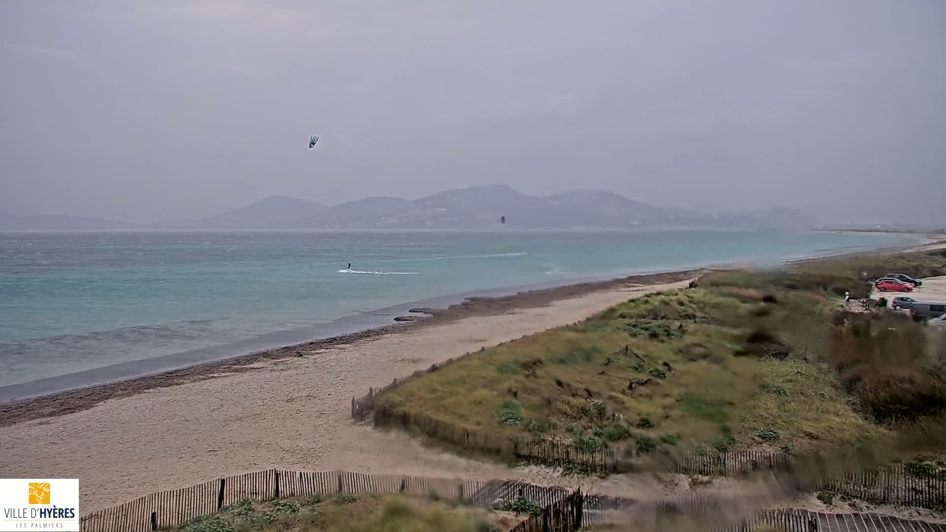 A sandy beach with a few kiteboarders in the water, under an overcast sky, with mountains visible in the distance.