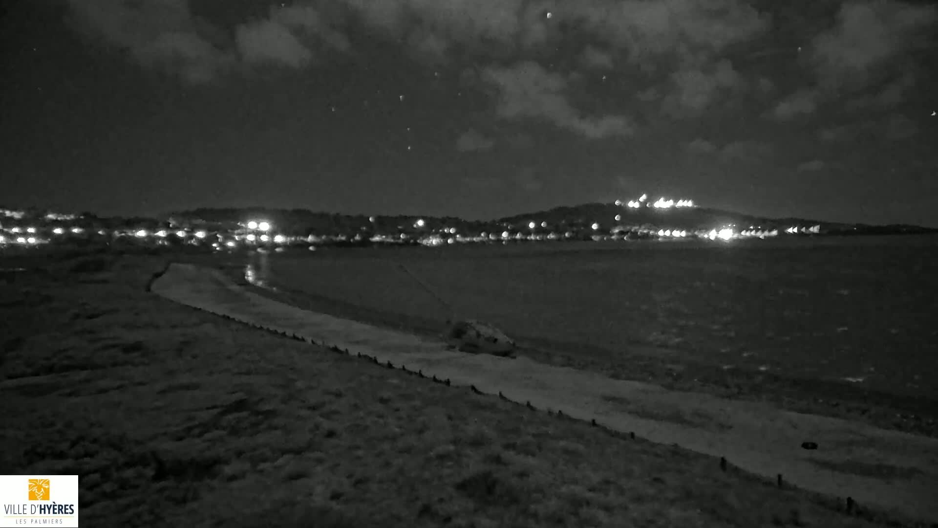 A nighttime, low-light view shows a body of water with a town's lights visible on the far shore, under a partly cloudy sky.