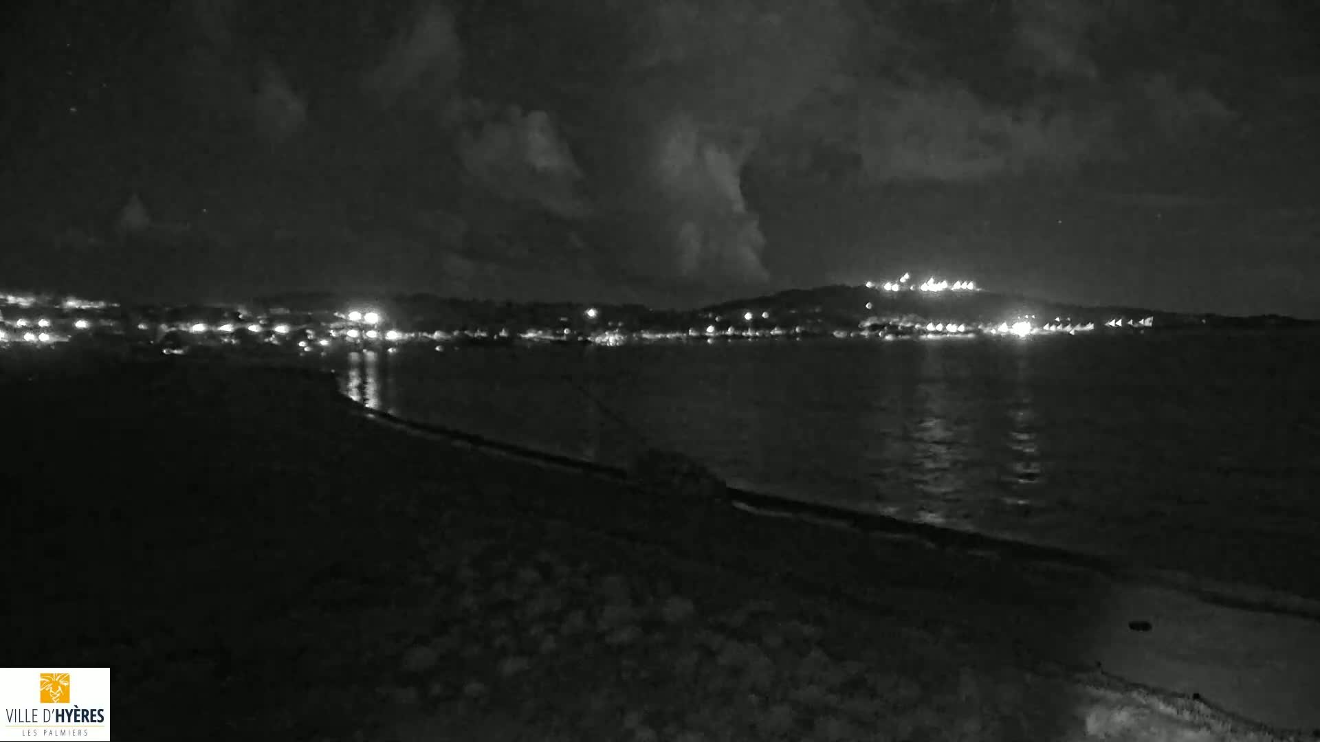 A nighttime view of a calm body of water reflecting the lights of a town on the opposite shore under a cloudy sky.