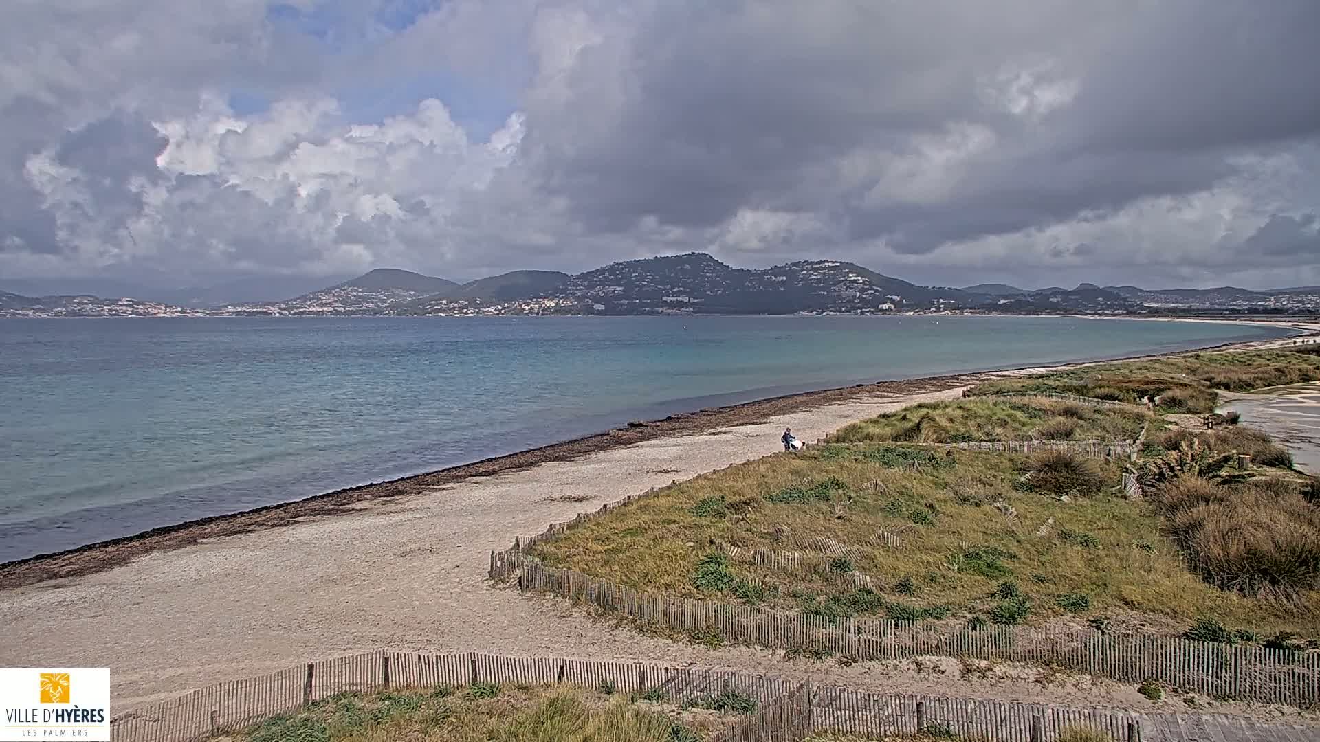 A sandy beach with a wooden fence borders a calm, teal-colored sea under a partly cloudy sky, with distant mountains visible in the background.