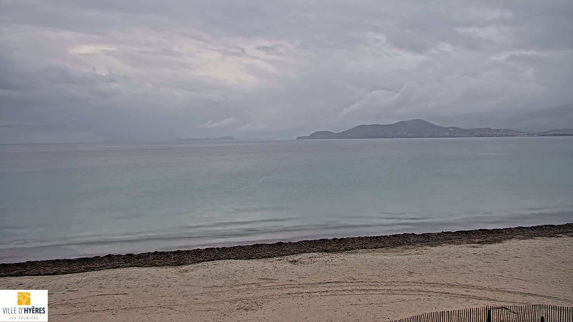 A sandy beach with a dark strip of seaweed at the water's edge faces a calm sea under a cloudy sky, with distant hills visible on the horizon.
