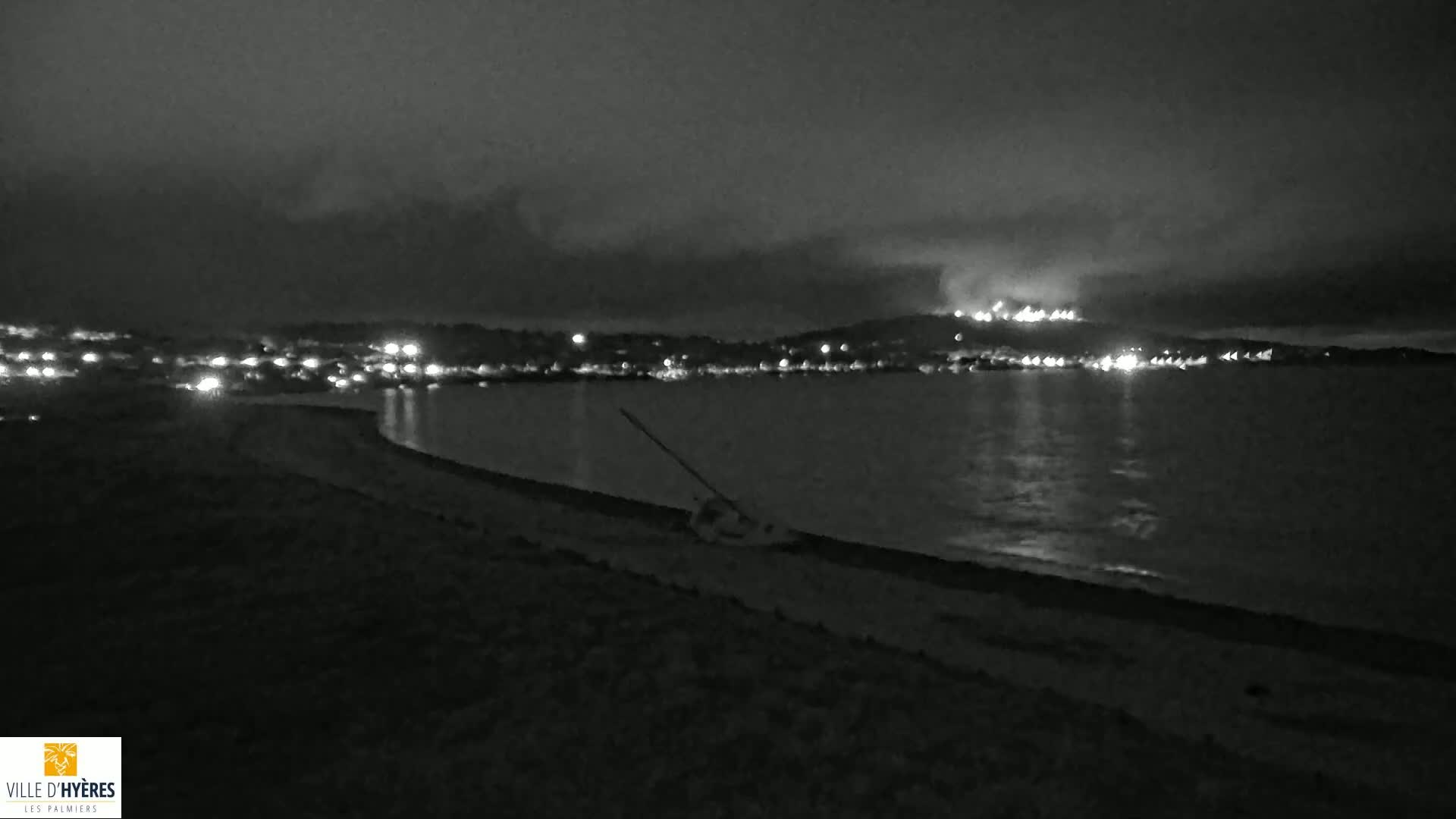 A dark, nighttime beach scene shows a body of water reflecting distant lights under a cloudy sky, with a piece of equipment lying on the sand.