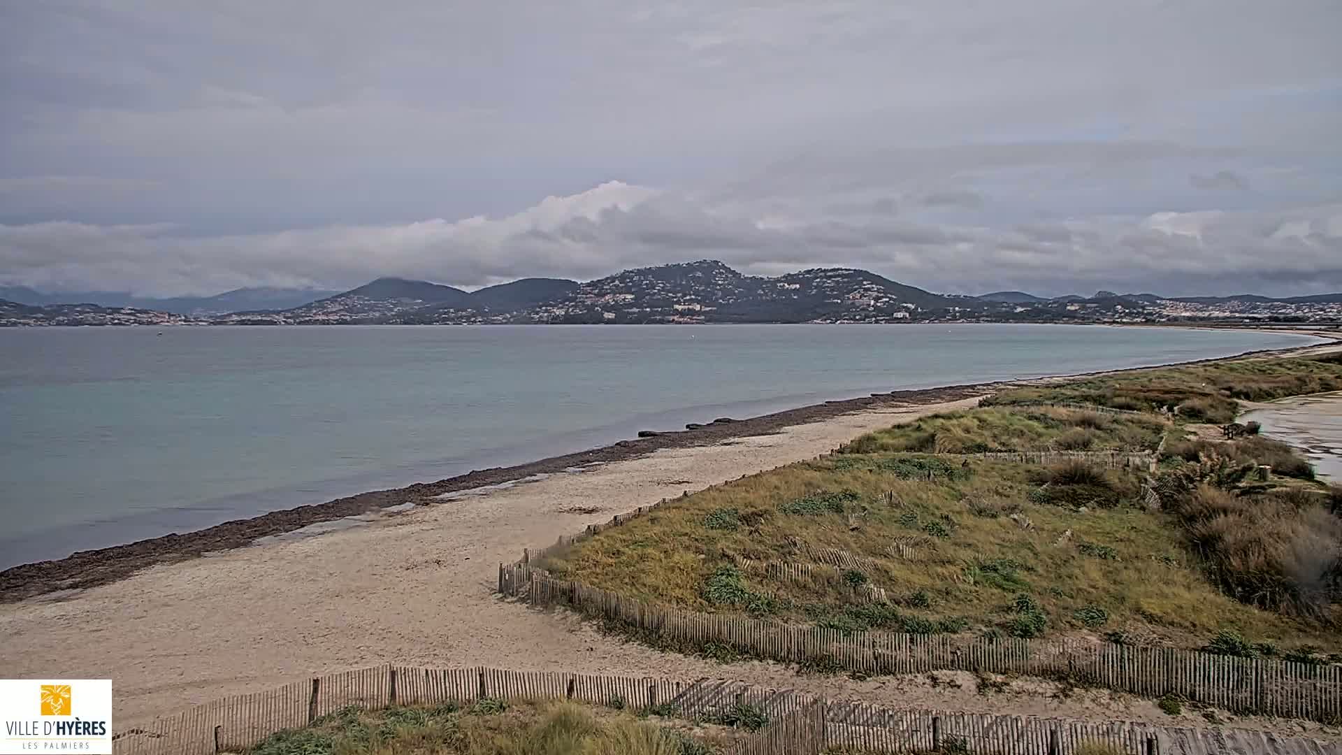 A sandy beach with low dunes and a wooden fence stretches along a calm, turquoise bay under a cloudy sky, with mountains visible in the distance.