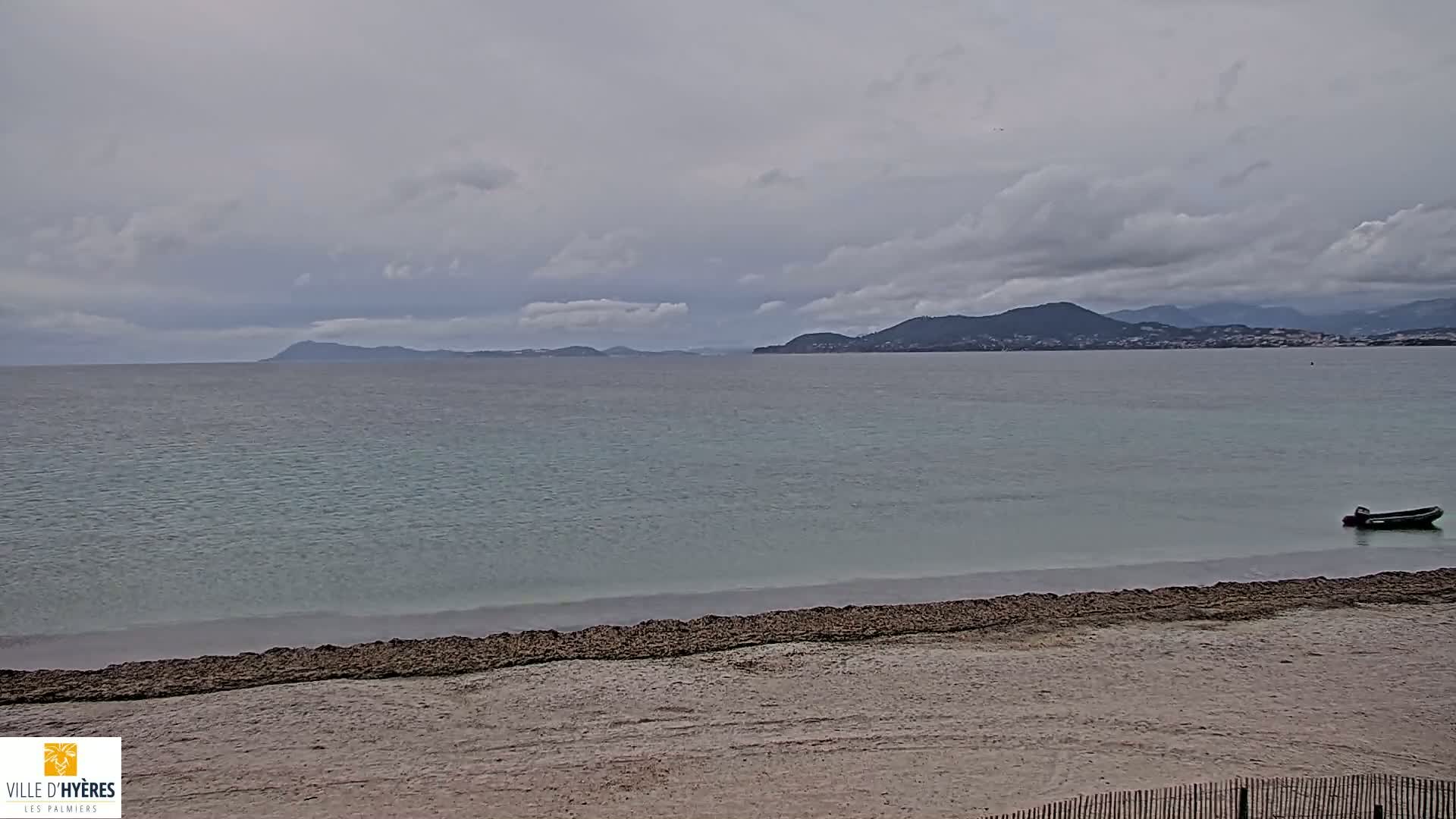 A sandy beach stretches to a calm, light-green sea under a cloudy sky, with distant mountains and a small boat visible in the water.