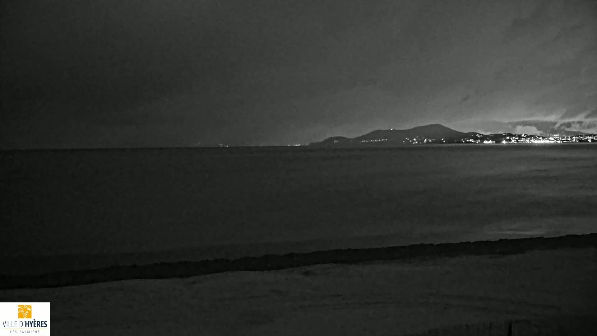 A dark, nighttime beach scene shows a calm sea and distant, dimly lit city lights on a hill.