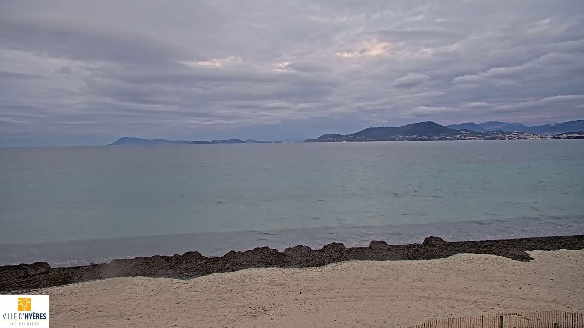 A sandy beach with dark rocks separating the sand from a calm, light-blue sea under a cloudy sky, with distant mountains visible on the horizon.