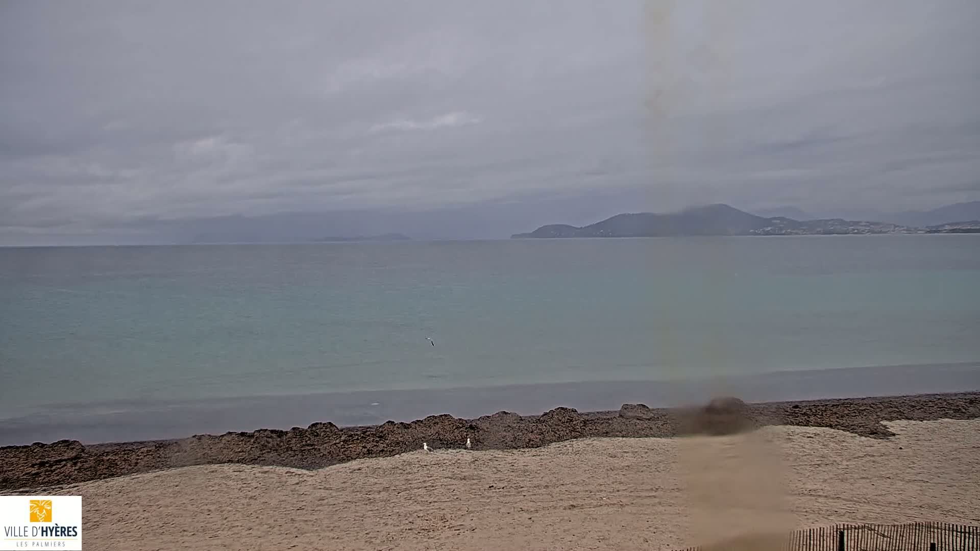 A sandy beach with dark seaweed at the waterline stretches toward a calm, light-blue sea under a cloudy, overcast sky, with distant hills visible on the horizon.