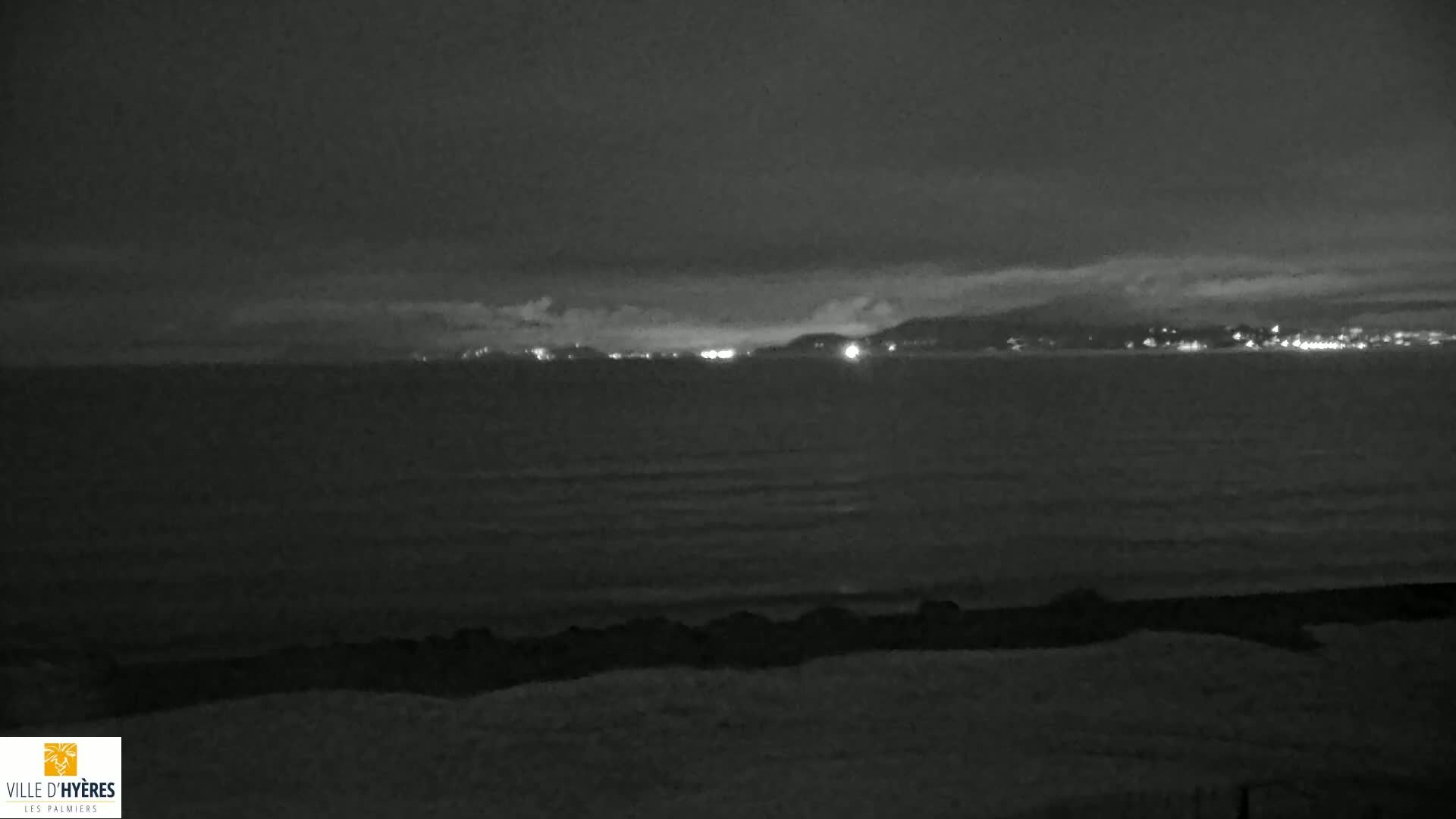 A nighttime, dark grayscale view of a calm ocean with a distant city skyline faintly illuminated.