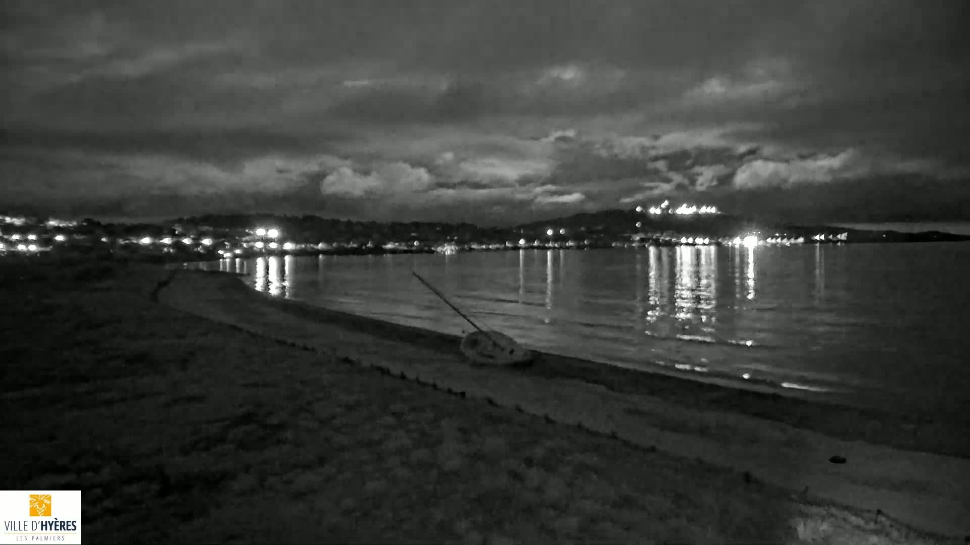 A nighttime, black and white view of a dark sandy beach with a small overturned sailboat on the shore, a calm sea reflecting city lights across the water under a cloudy sky.