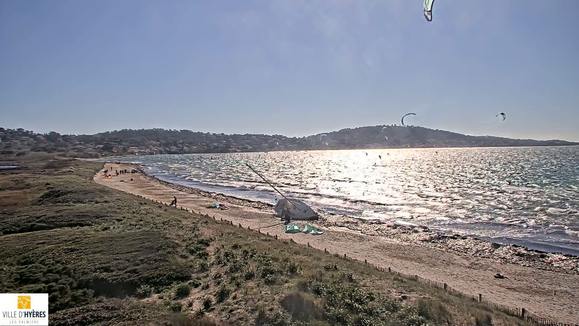 A sandy beach with several people and kitesurfers is visible, with a calm, sunlit sea and distant hills under a clear sky.
