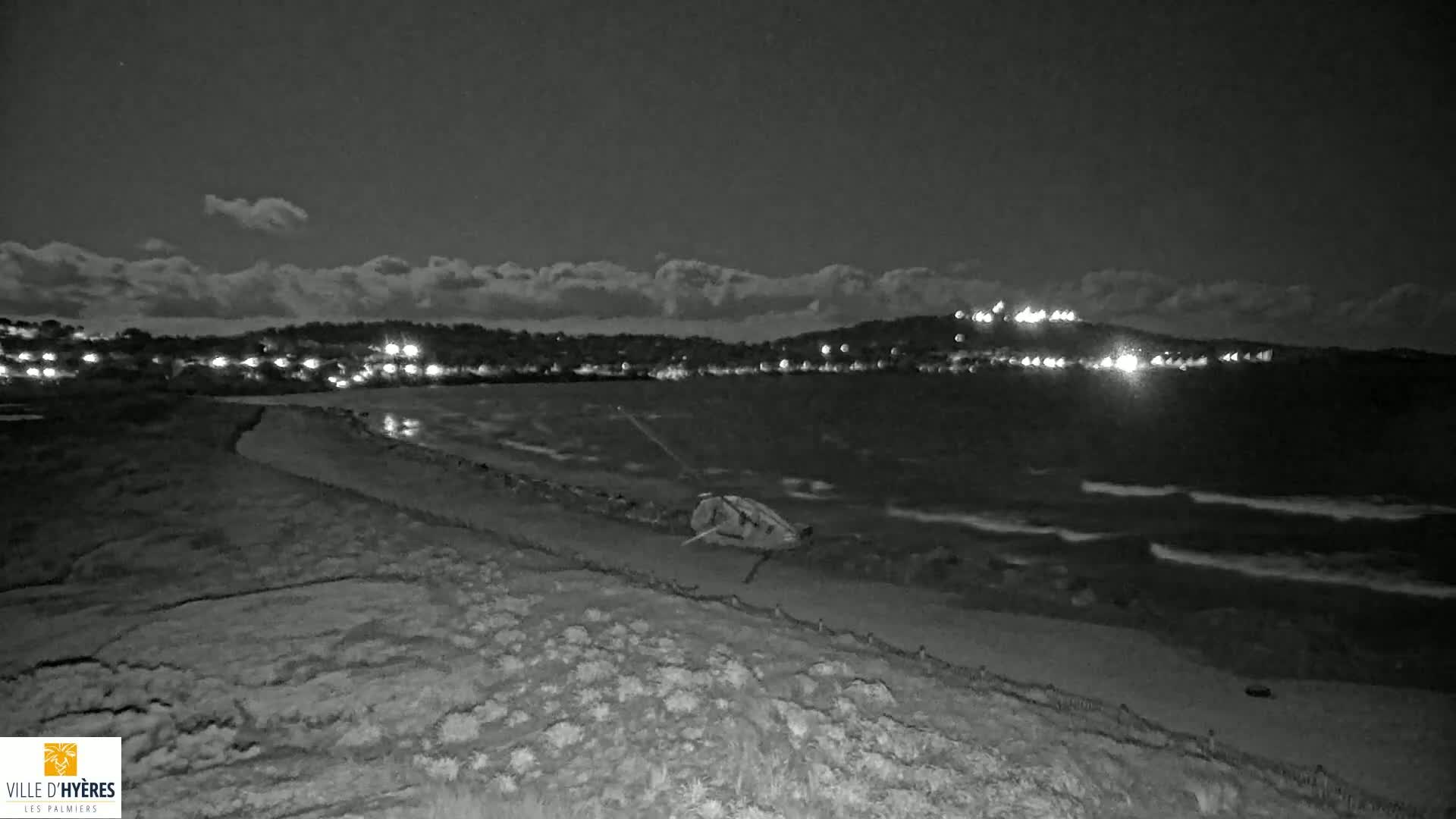 A nighttime, black and white view of a wavy ocean under a partly cloudy sky, with lights visible on the far shore and an object resembling a small boat on the beach in the foreground.