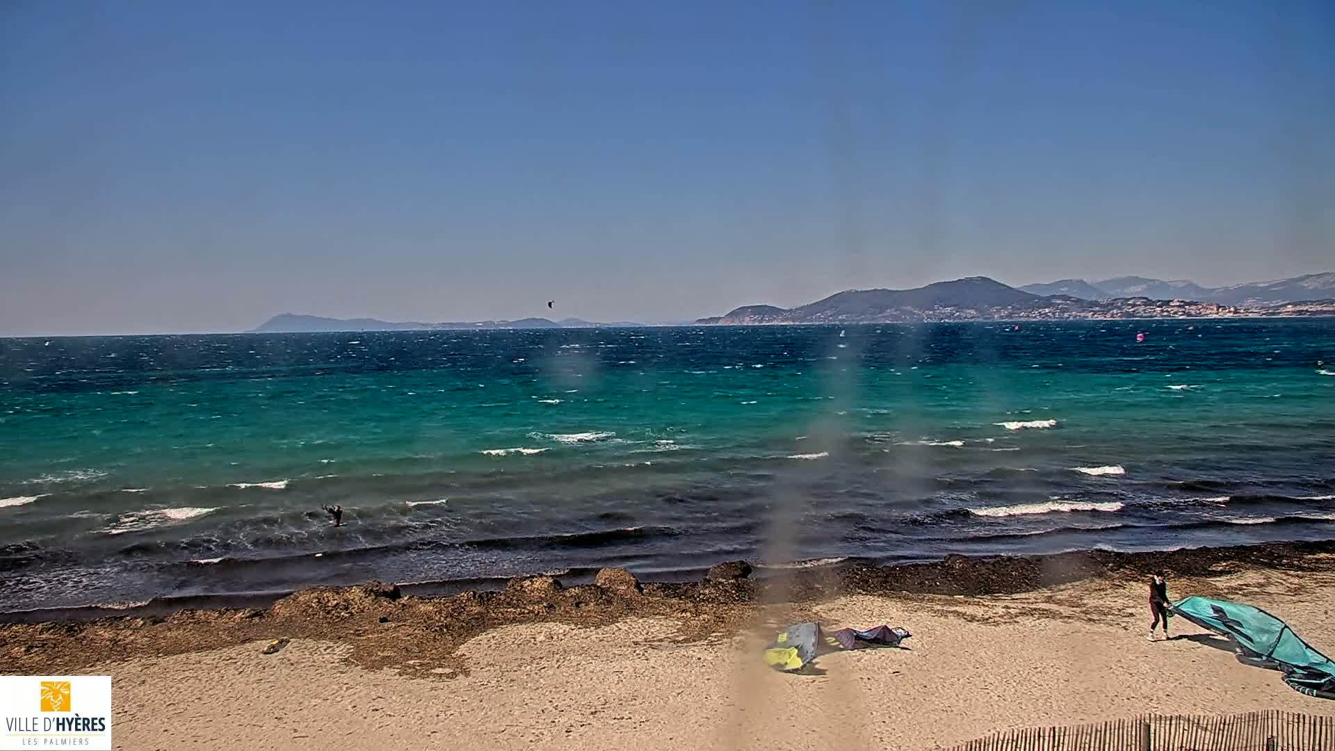 A windy, sunny day at a beach shows turquoise water with small waves, several kitesurfers in the distance, and some kitesurfers on the sandy shore.