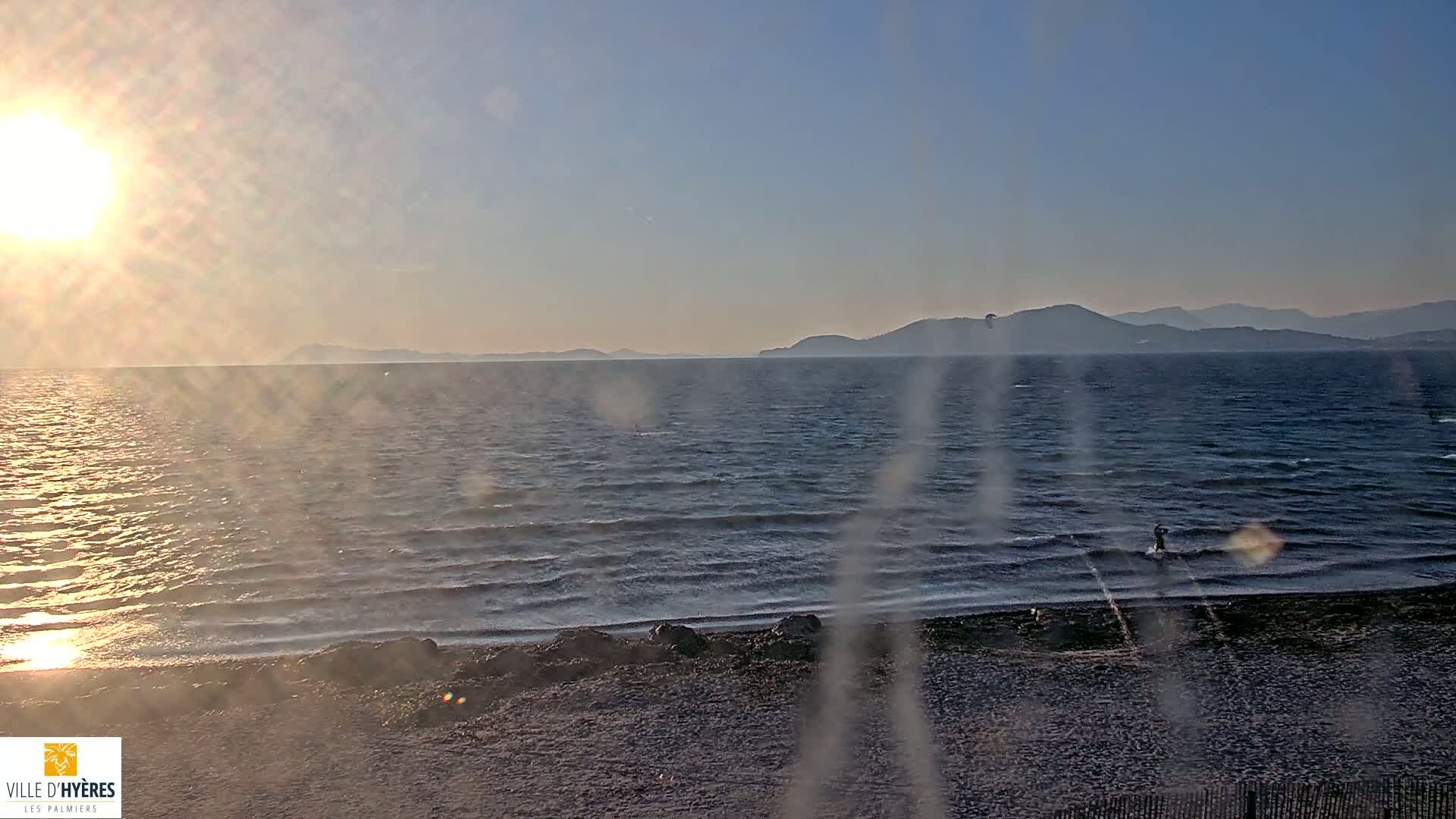 A person kitesurfs in a wavy ocean near a beach at sunset, with mountains visible in the distance.