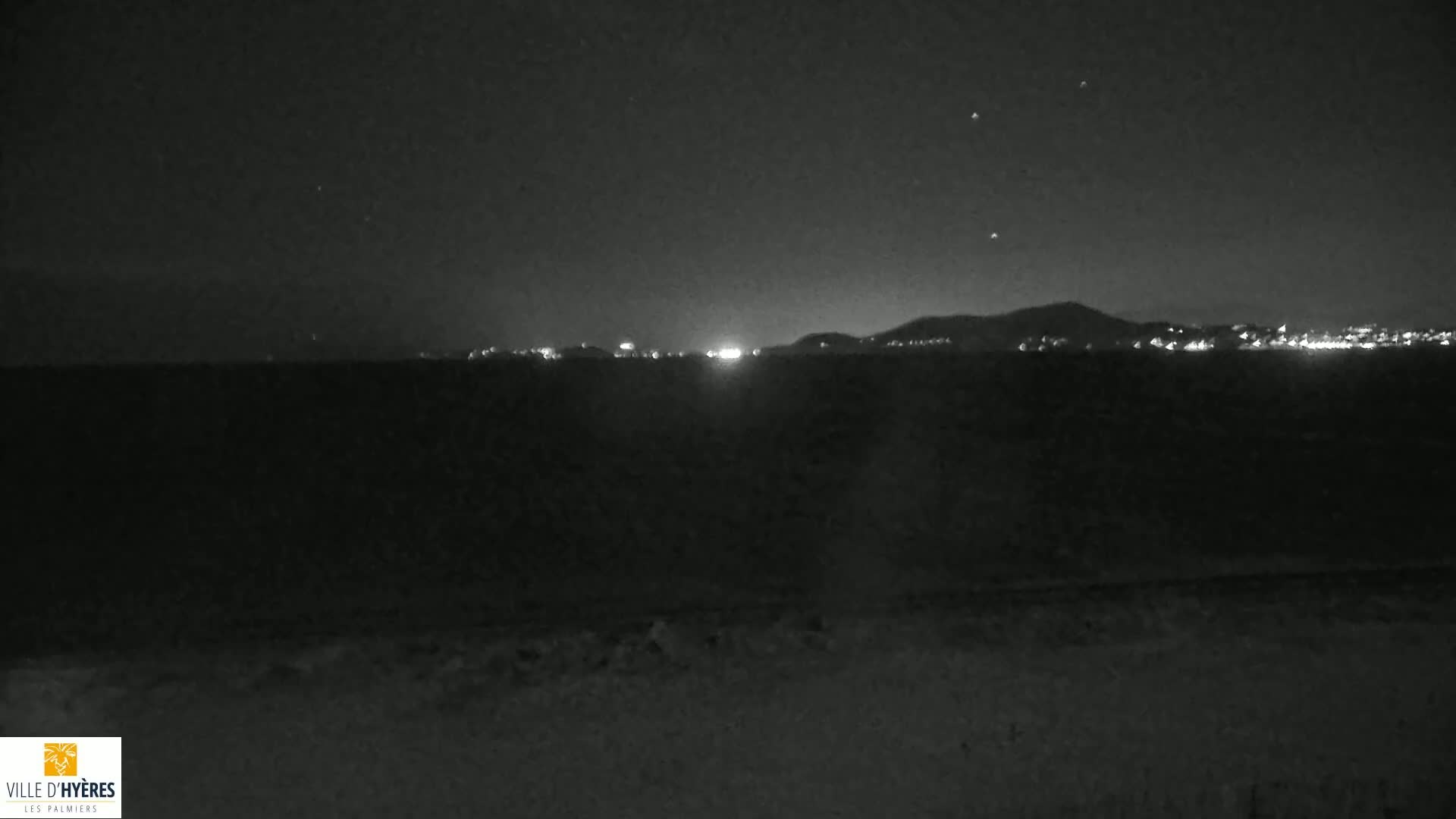 A nighttime view of a dark shoreline with a distant city glowing under a clear, starlit sky.