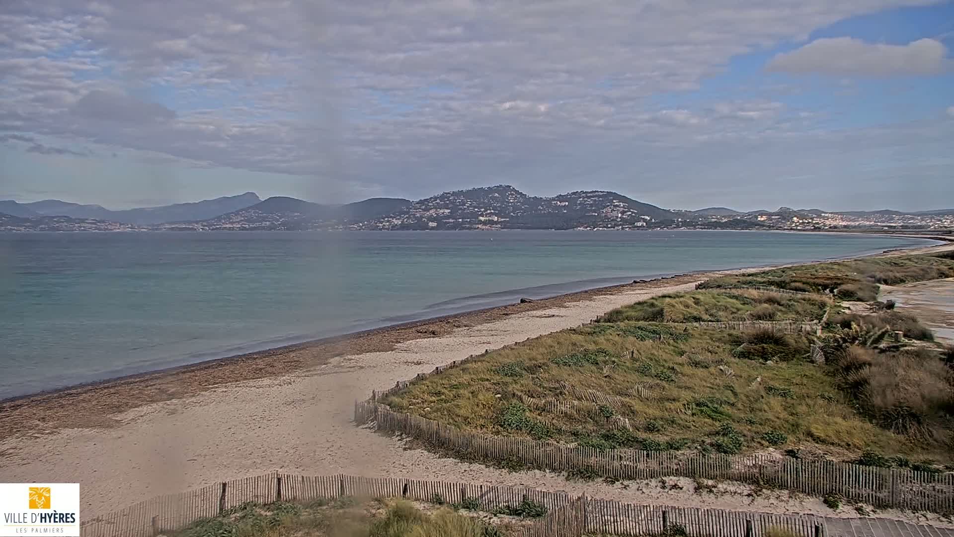 A sandy beach with low dunes and a wooden fence stretches along a calm, teal-colored sea under a cloudy sky, with distant mountains visible on the horizon.