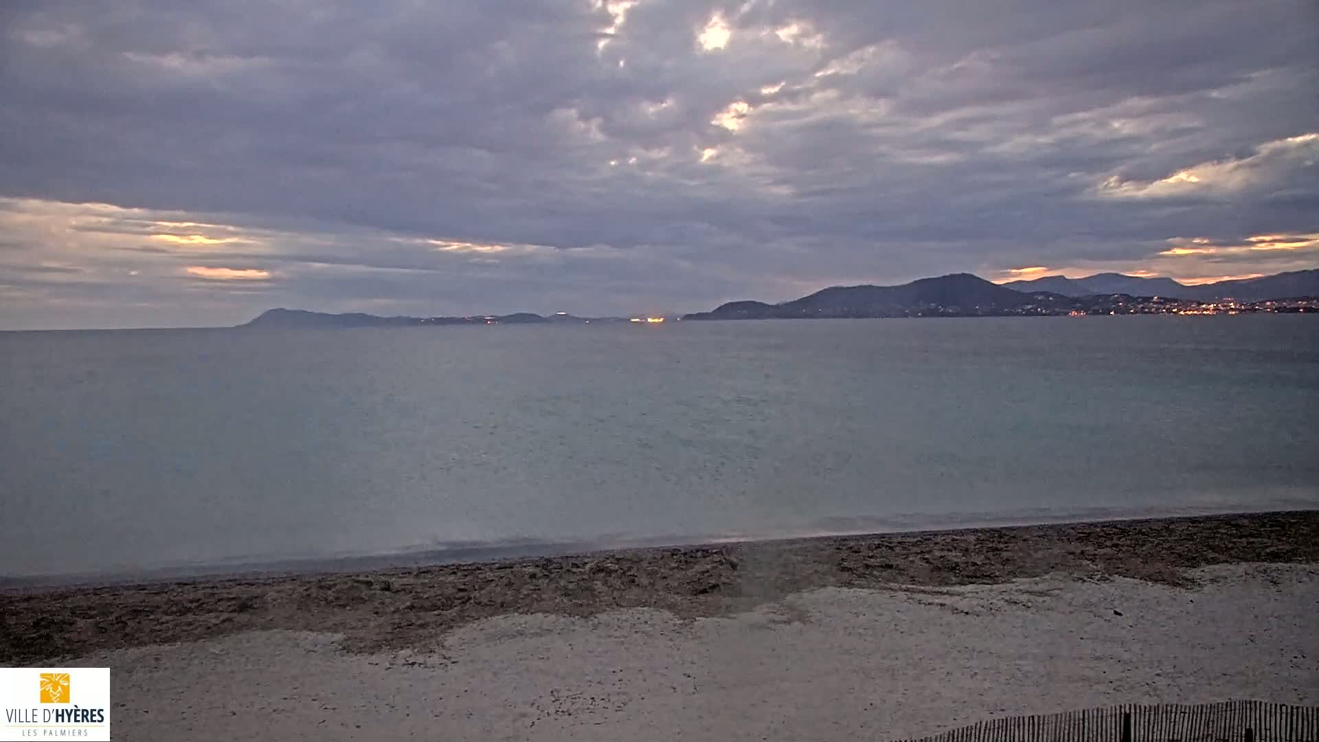 A sandy beach meets a calm sea under a cloudy, dusky sky, with distant mountains and lights visible on the horizon.