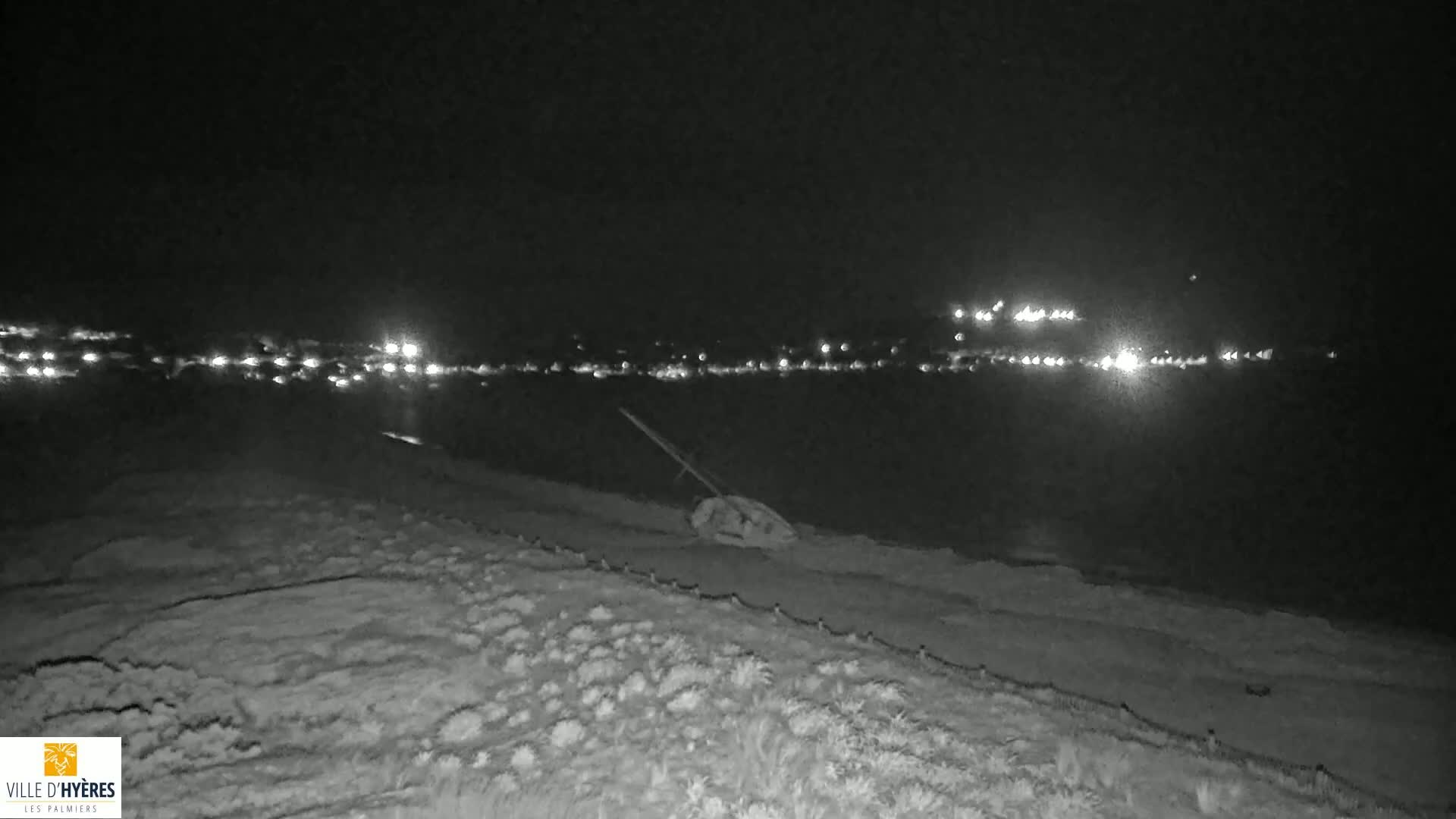 A nighttime, low-light view of a shoreline with distant city lights across a body of water, and a piece of debris near the foreground.