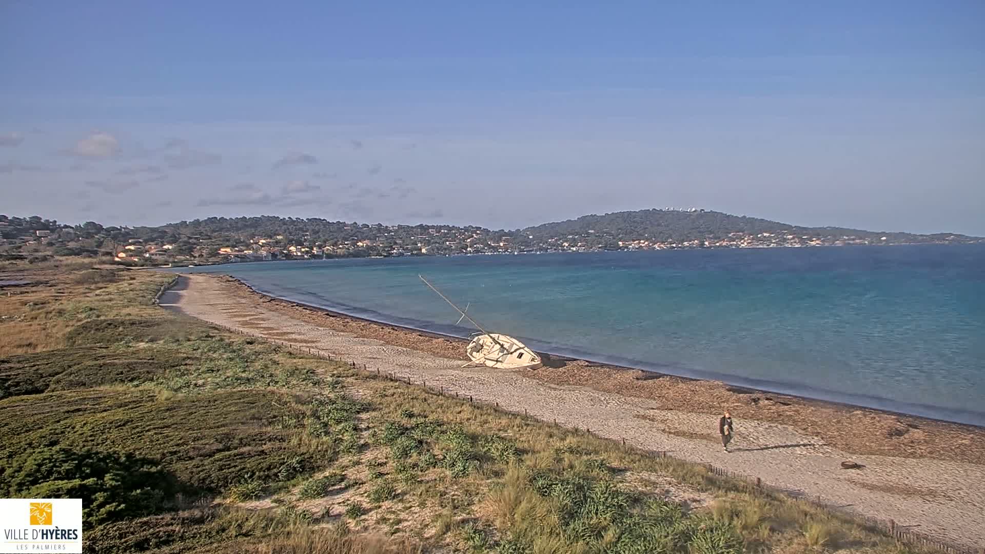 A sandy beach with a capsized sailboat and a person walking along the shore, overlooks a calm, turquoise sea and distant hills under a clear blue sky.