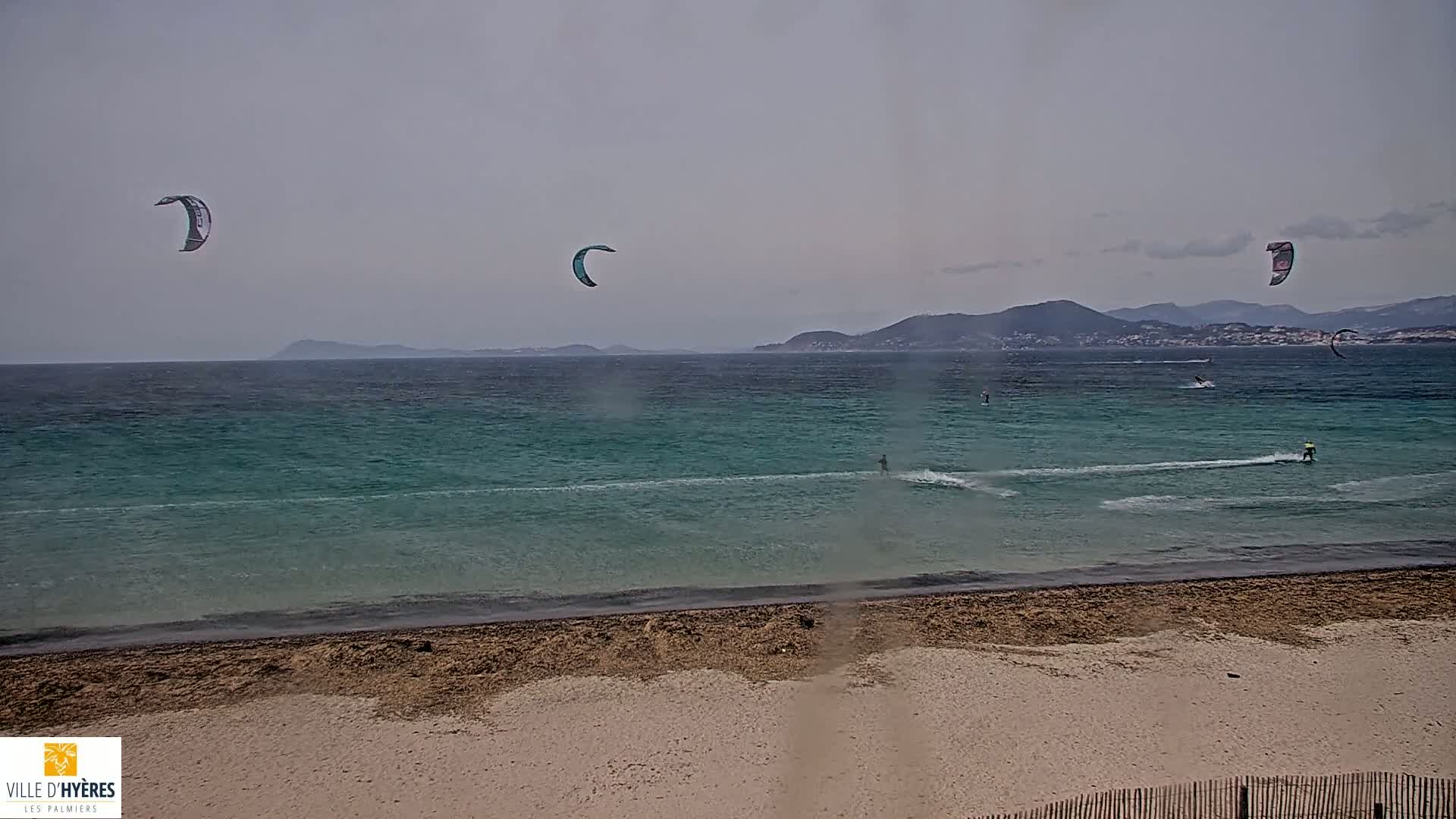 Several kitesurfers ride in a teal ocean under an overcast sky on a sandy beach.