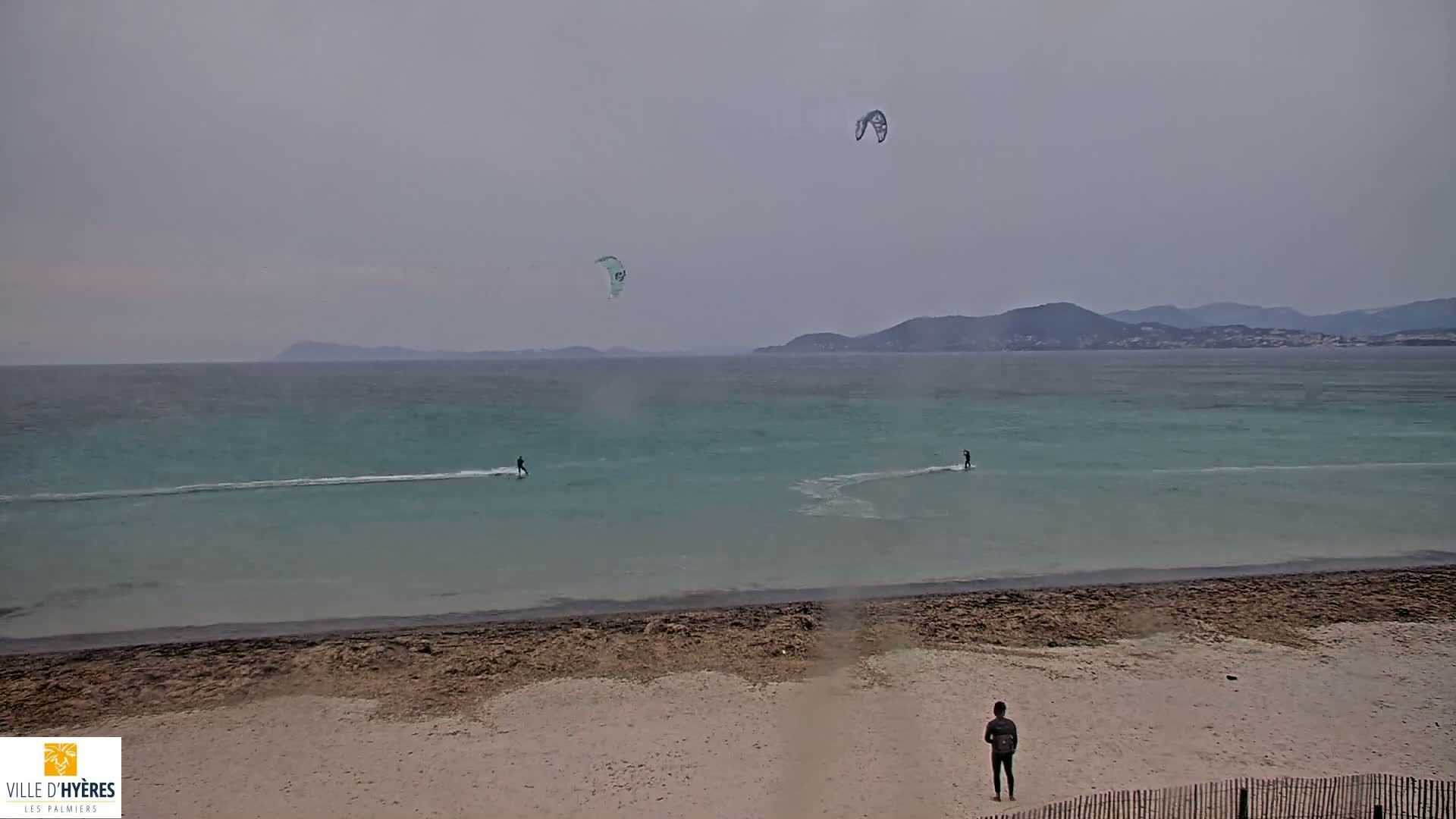 Two kitesurfers ride the turquoise water of a calm sea under an overcast sky, while a third person watches from a sandy beach.