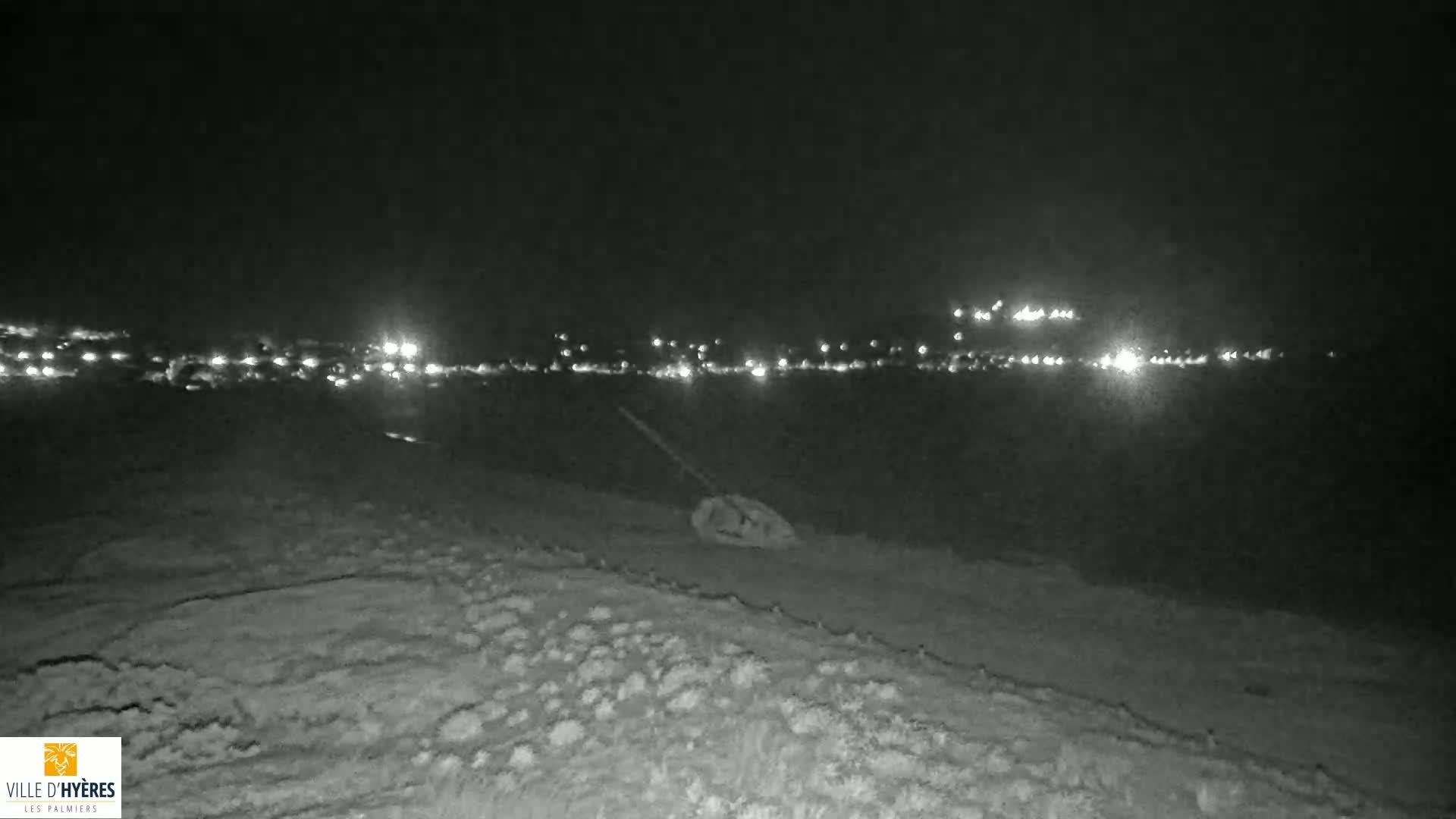 A nighttime view shows a distant line of lights across a dark body of water, with a rough, textured foreground under a clear, dark sky.