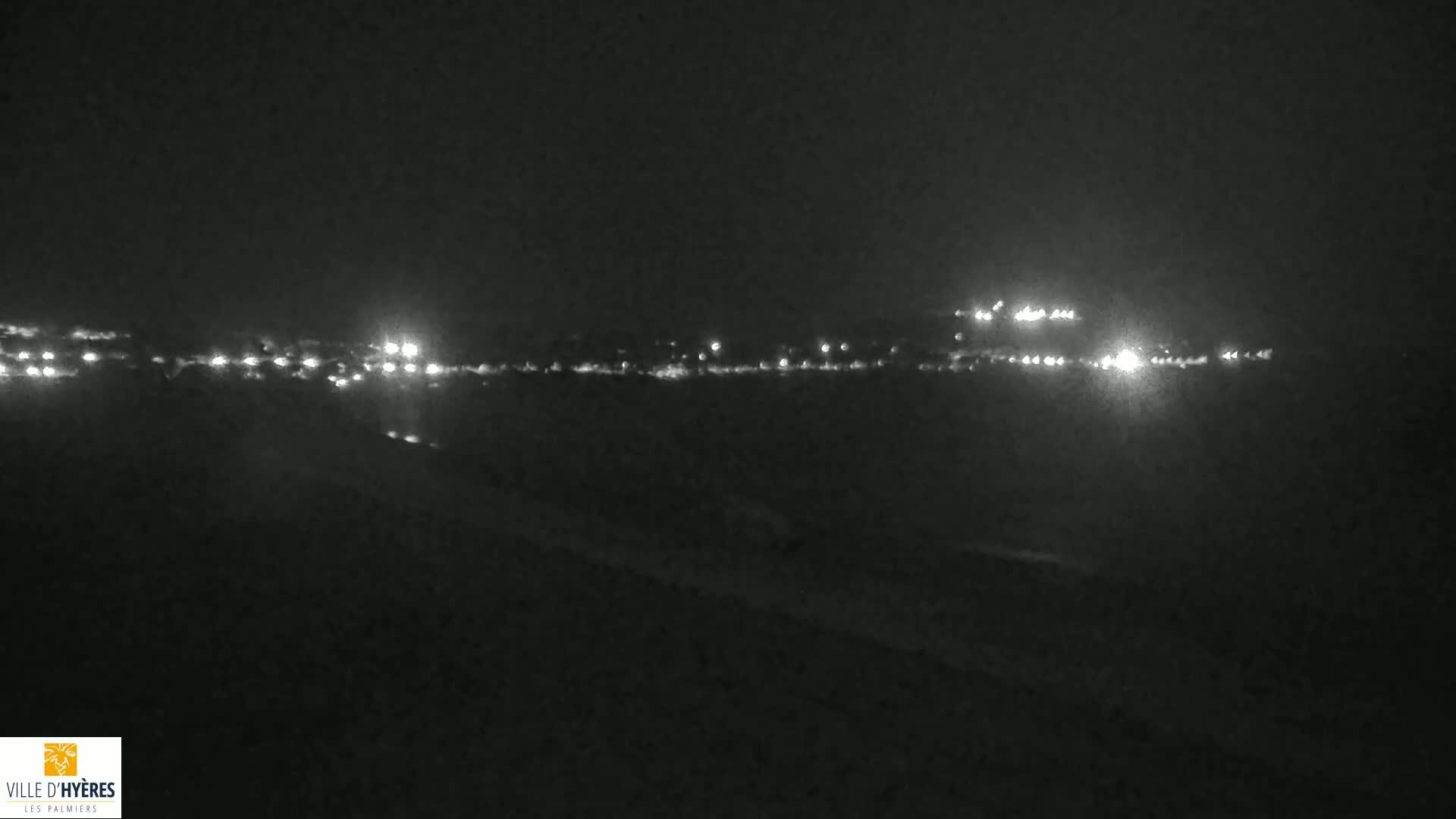 A nighttime long shot shows a distant line of city lights across a dark landscape under a clear, dark sky.