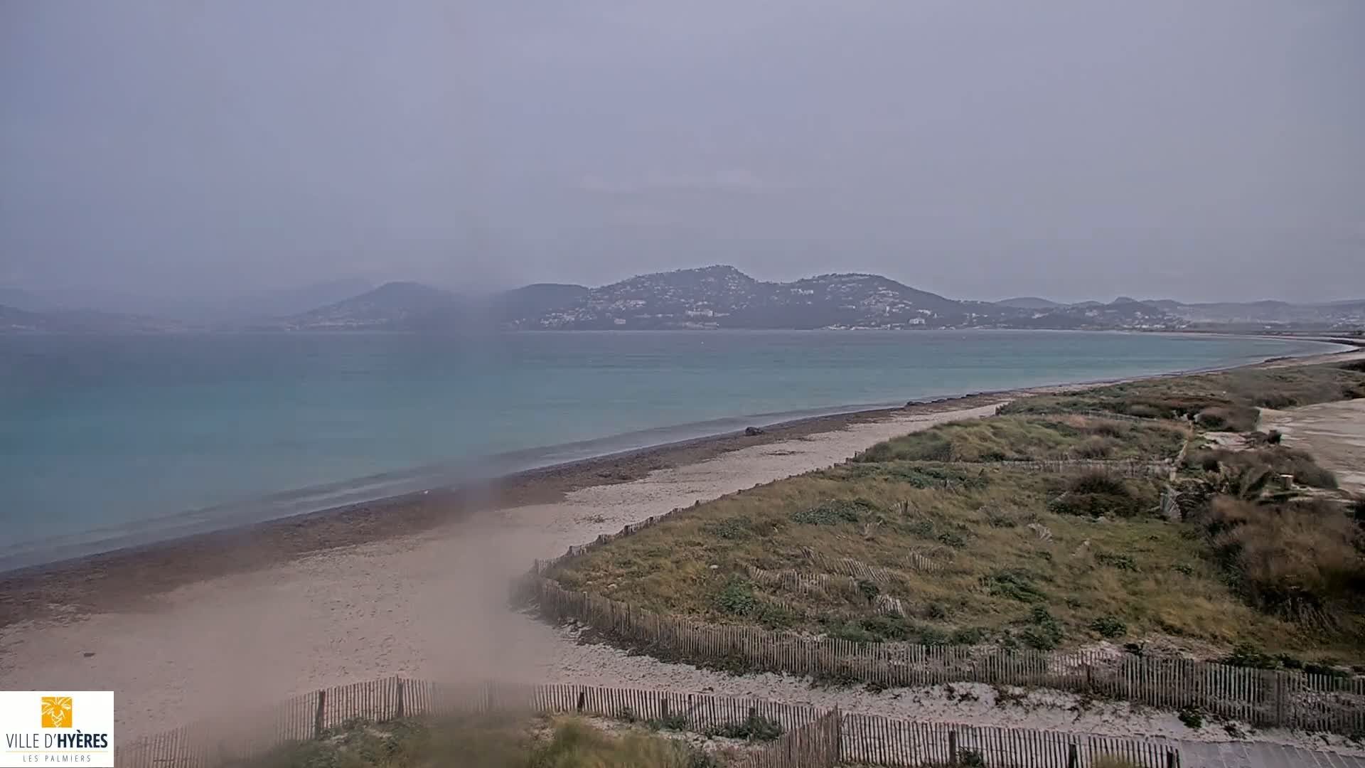 A sandy beach bordered by dunes and a wooden fence stretches along a calm, teal sea under an overcast sky.