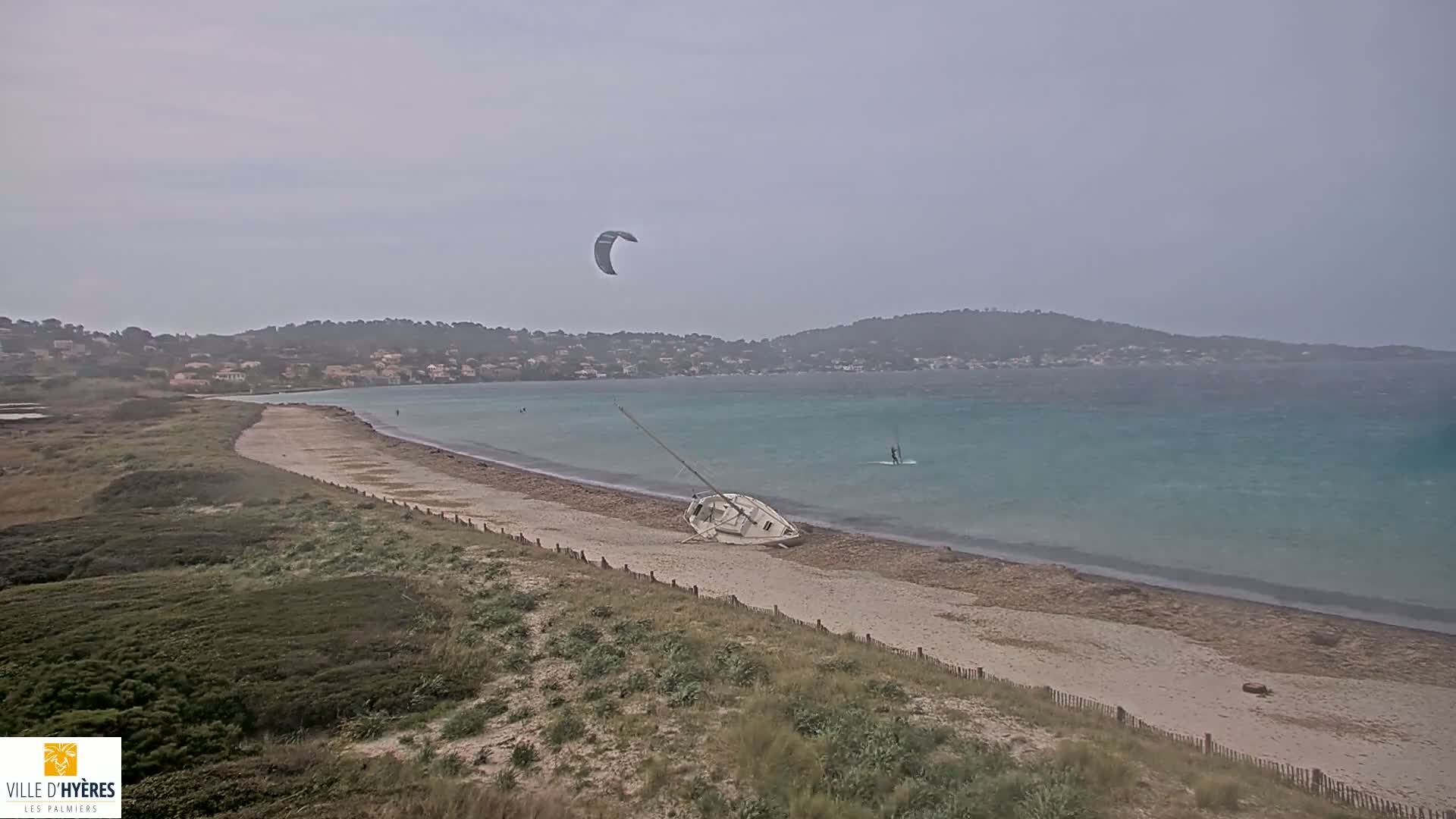 A partly cloudy day shows a sandy beach with a capsized sailboat, a kite surfer, and a wind surfer on a turquoise sea, backed by a low, tree-covered hillside.