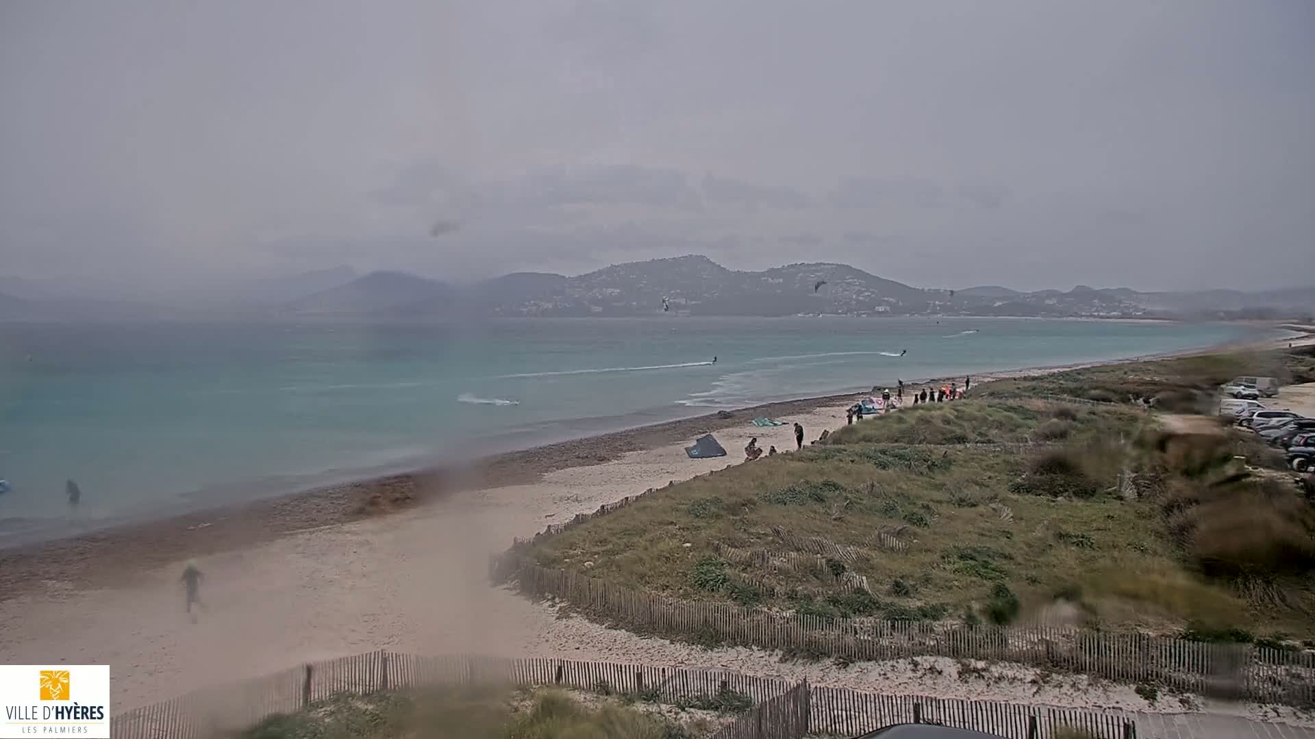 A beach with a few people on it and several kitesurfers in the water, under an overcast sky.