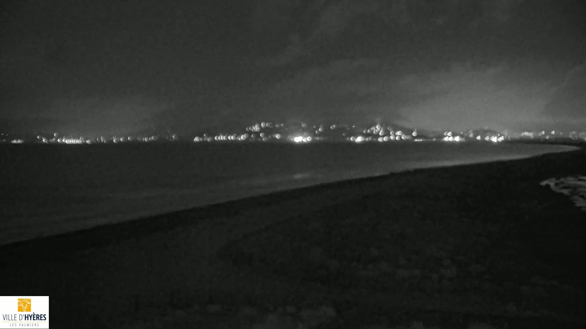 A dark, nighttime beach scene shows a distant city skyline illuminated with lights across a calm body of water under a cloudy sky.