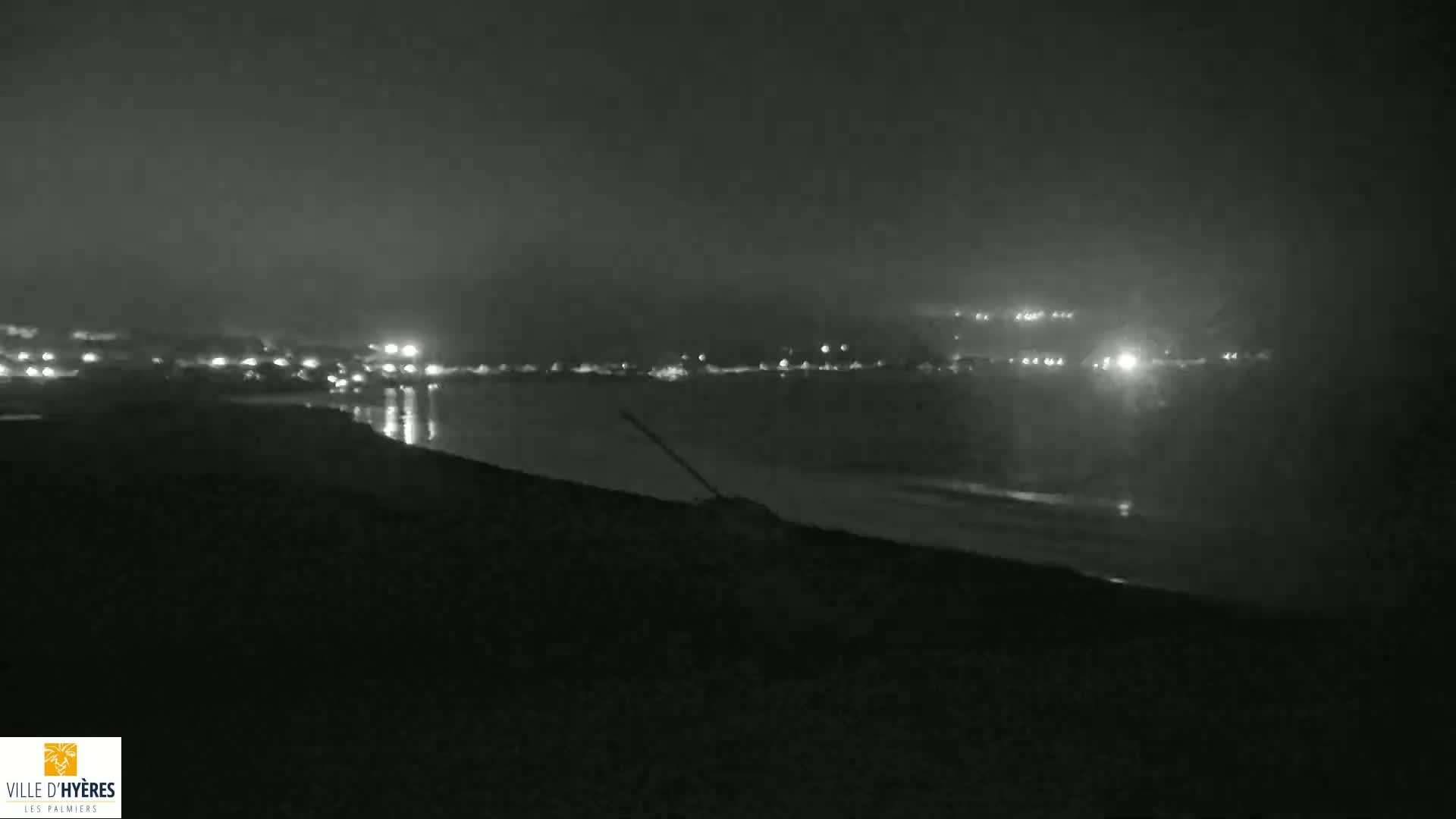 A dark, nighttime beach scene shows a body of water with distant lights reflecting on its surface under a hazy sky.