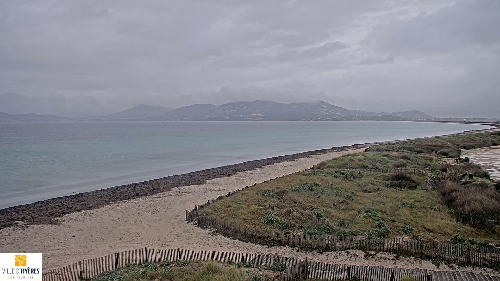 A sandy beach with dark seaweed along the water's edge curves gently around a bay under a cloudy, overcast sky, with distant mountains visible across the calm water.