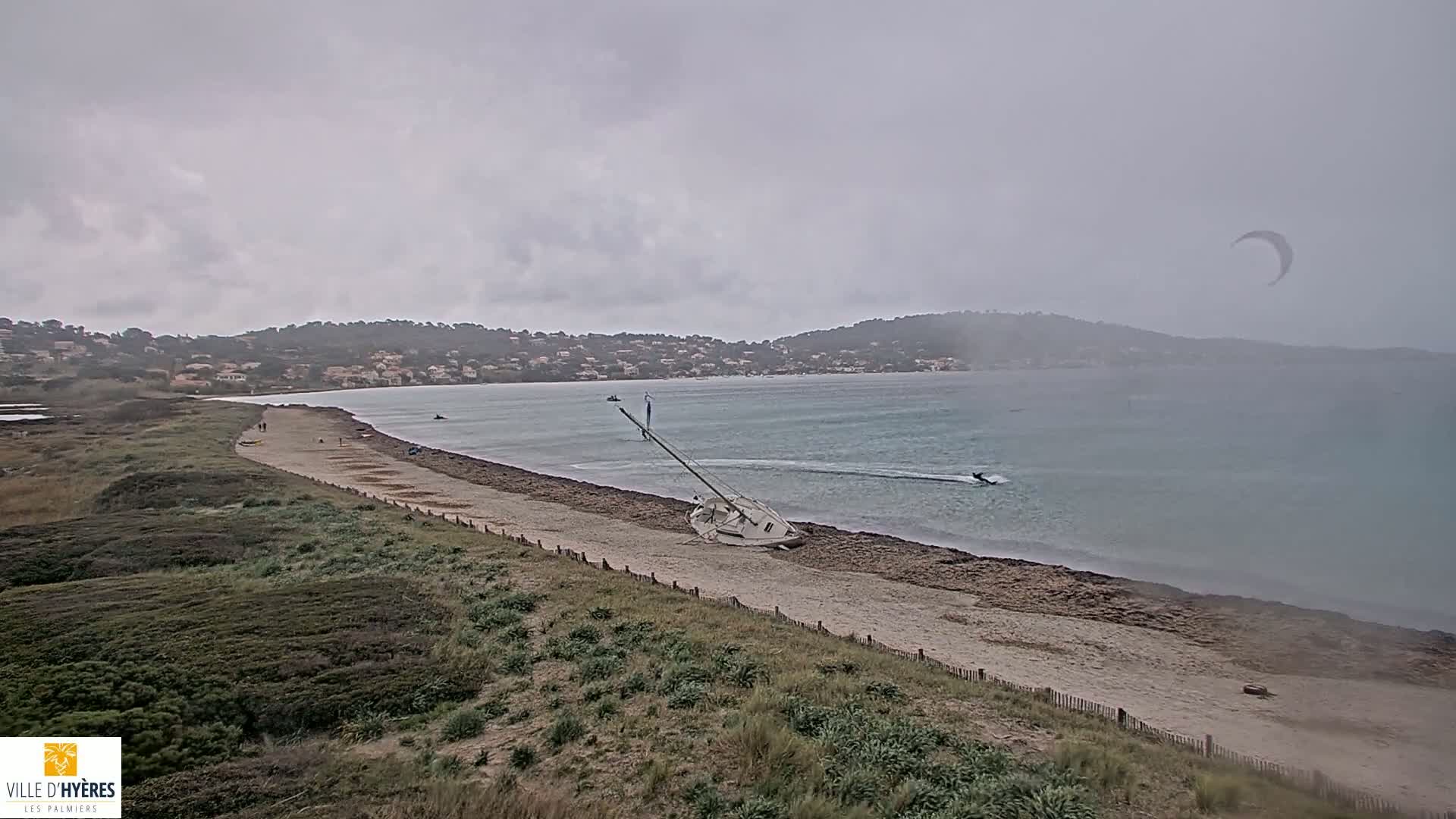 A sandy beach with a capsized sailboat and a kitesurfer in the water is visible under an overcast sky.