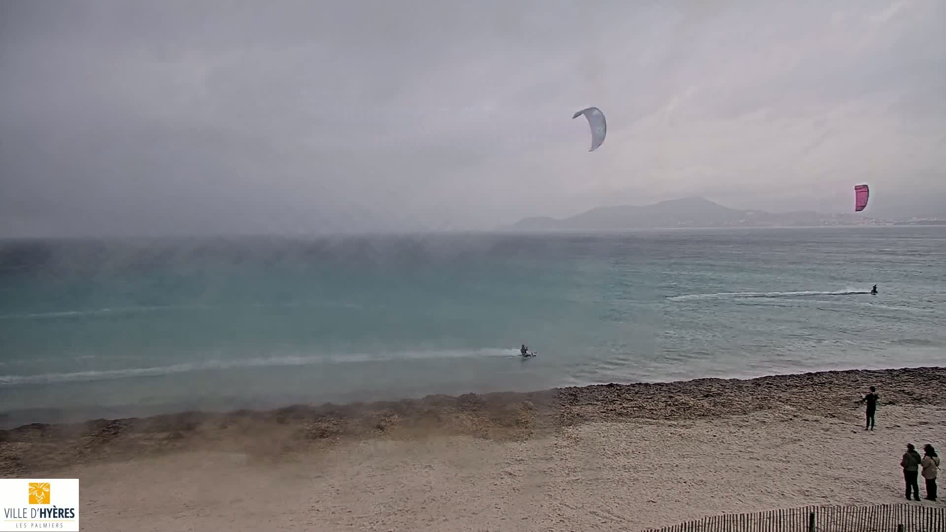 A sandy beach with a few people on it overlooks a calm sea where several kite surfers are riding in misty, overcast conditions.