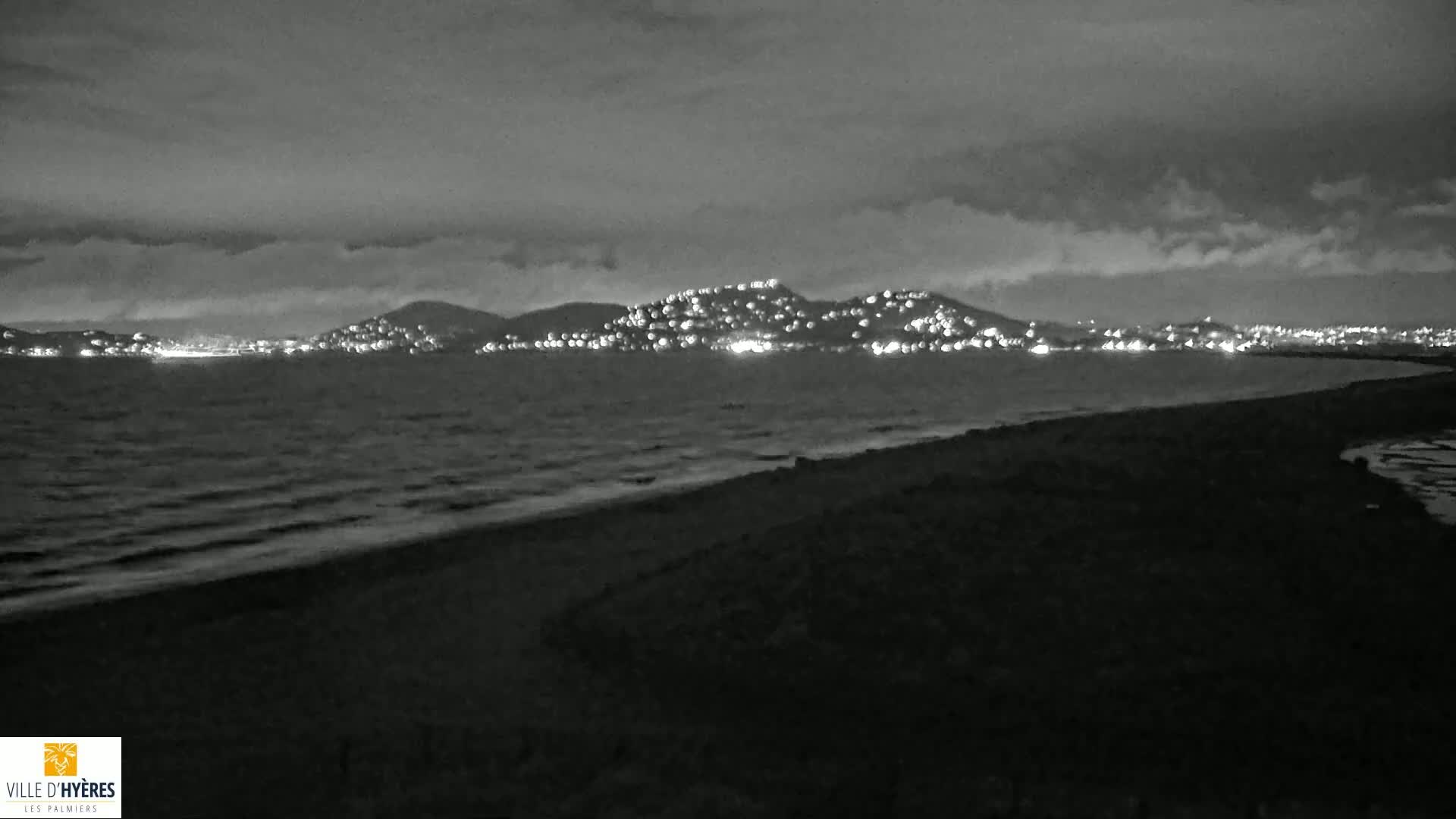 A nighttime, grayscale view of a dark beach and ocean with a distant, hilltop town illuminated by lights under a cloudy sky.